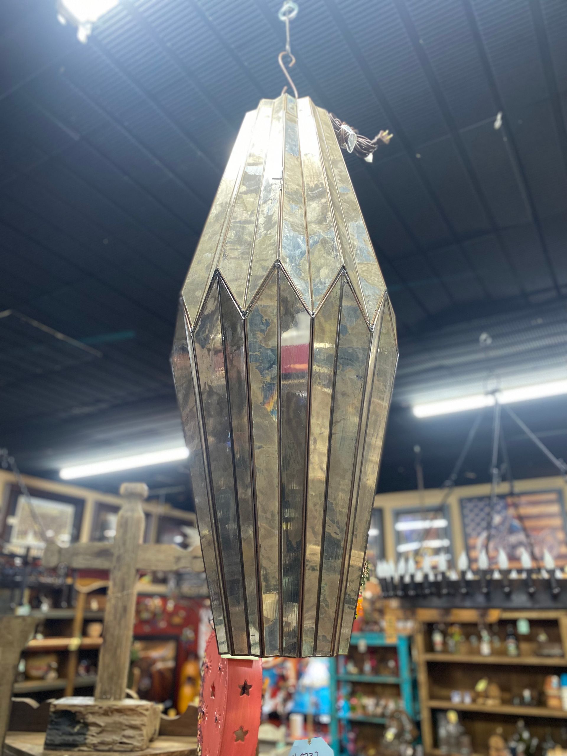 A silver, diamond-shaped hanging lantern with a decorative, geometric design, likely for indoor use. It is hanging in a shop with other items visible.