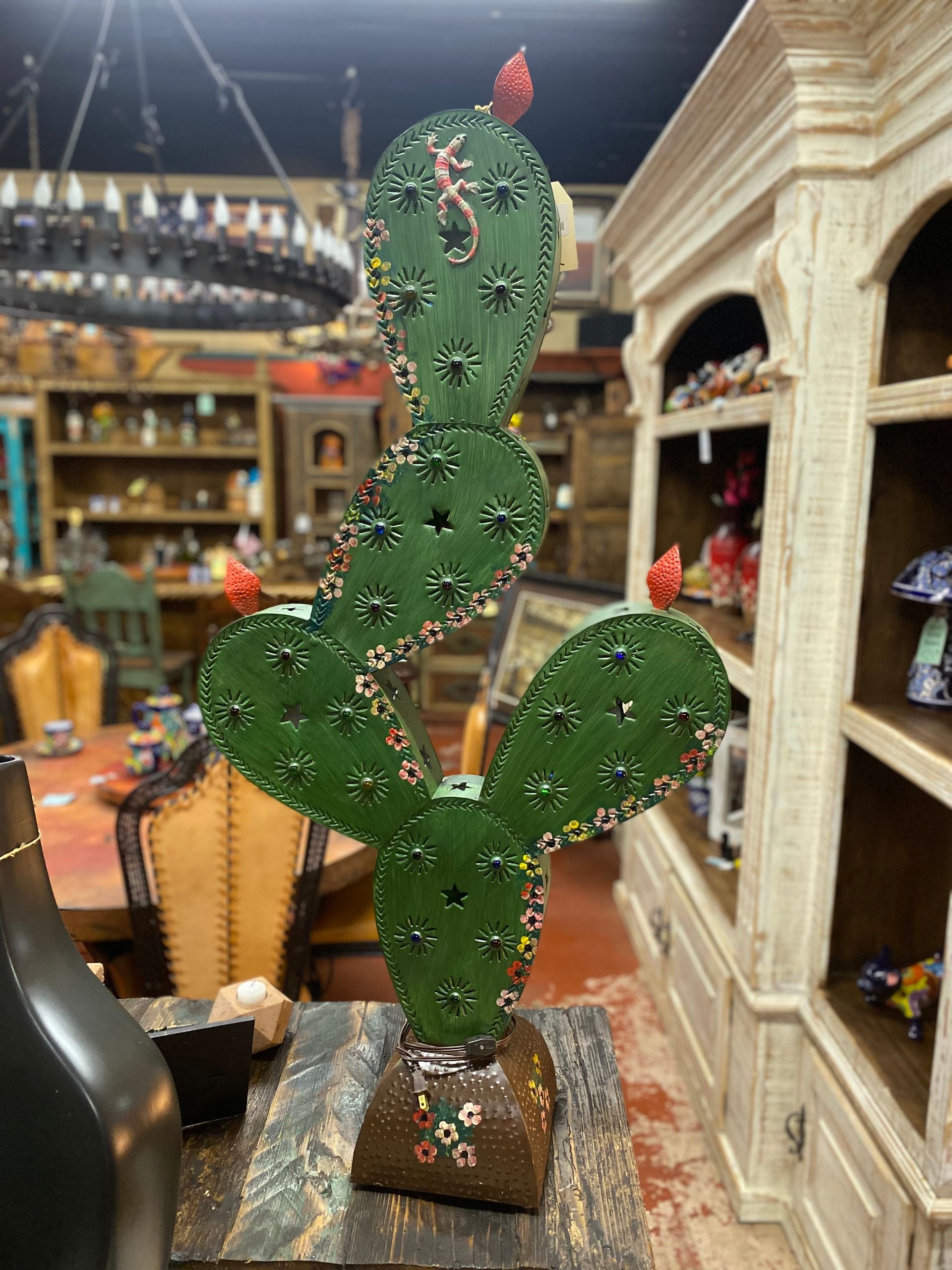 A decorative green metal cactus with colorful flower accents, displayed indoors on a wooden surface near a white cabinet.