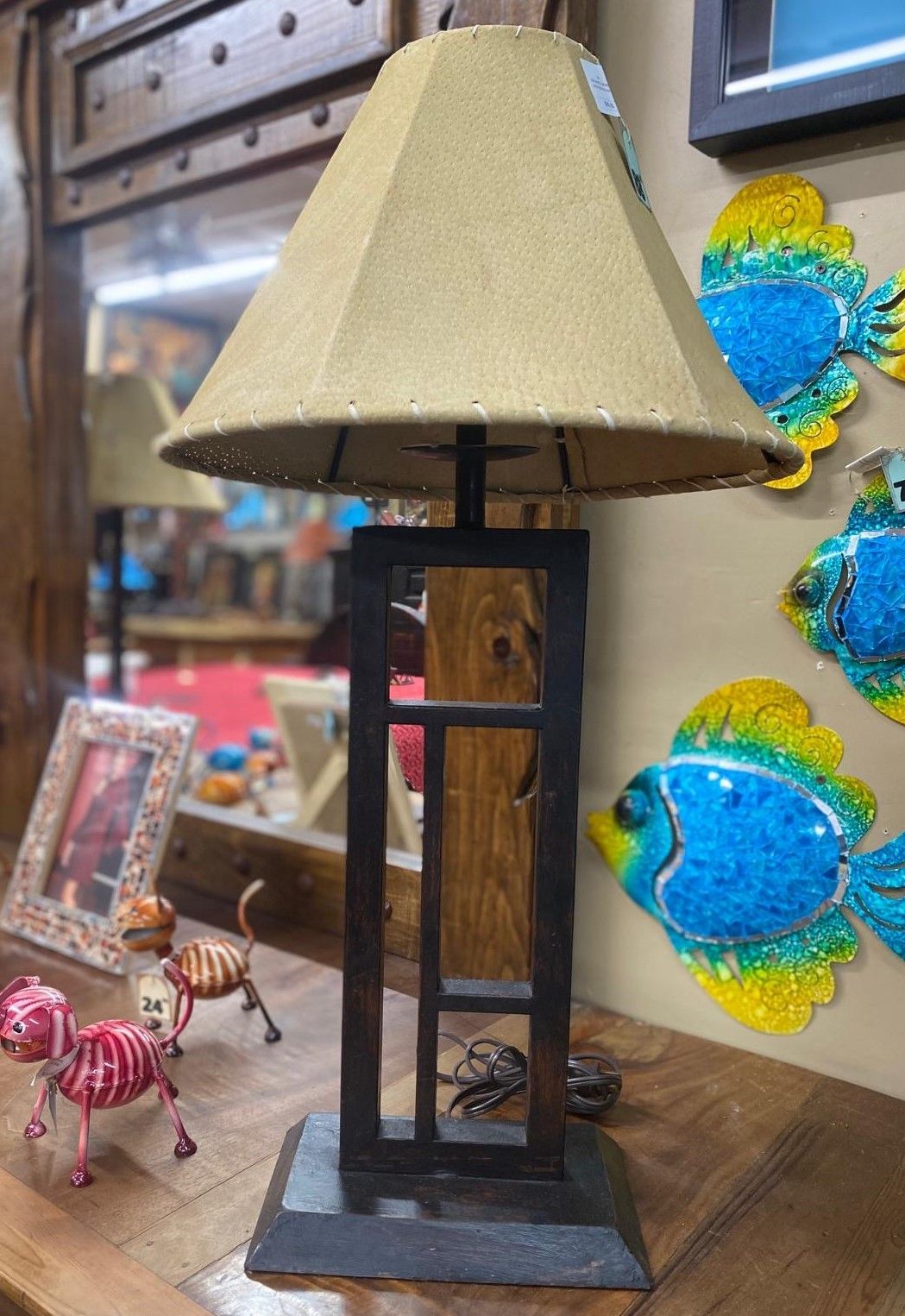 A tall, rectangular, black metal lamp with a textured tan lampshade sits on a wooden shelf. The background has decorative items and a mirror.