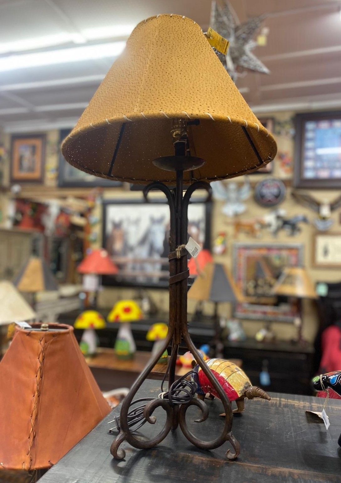 A rustic metal table lamp with a perforated tan lampshade. It sits on a wooden surface in a shop filled with other lamps and decor.