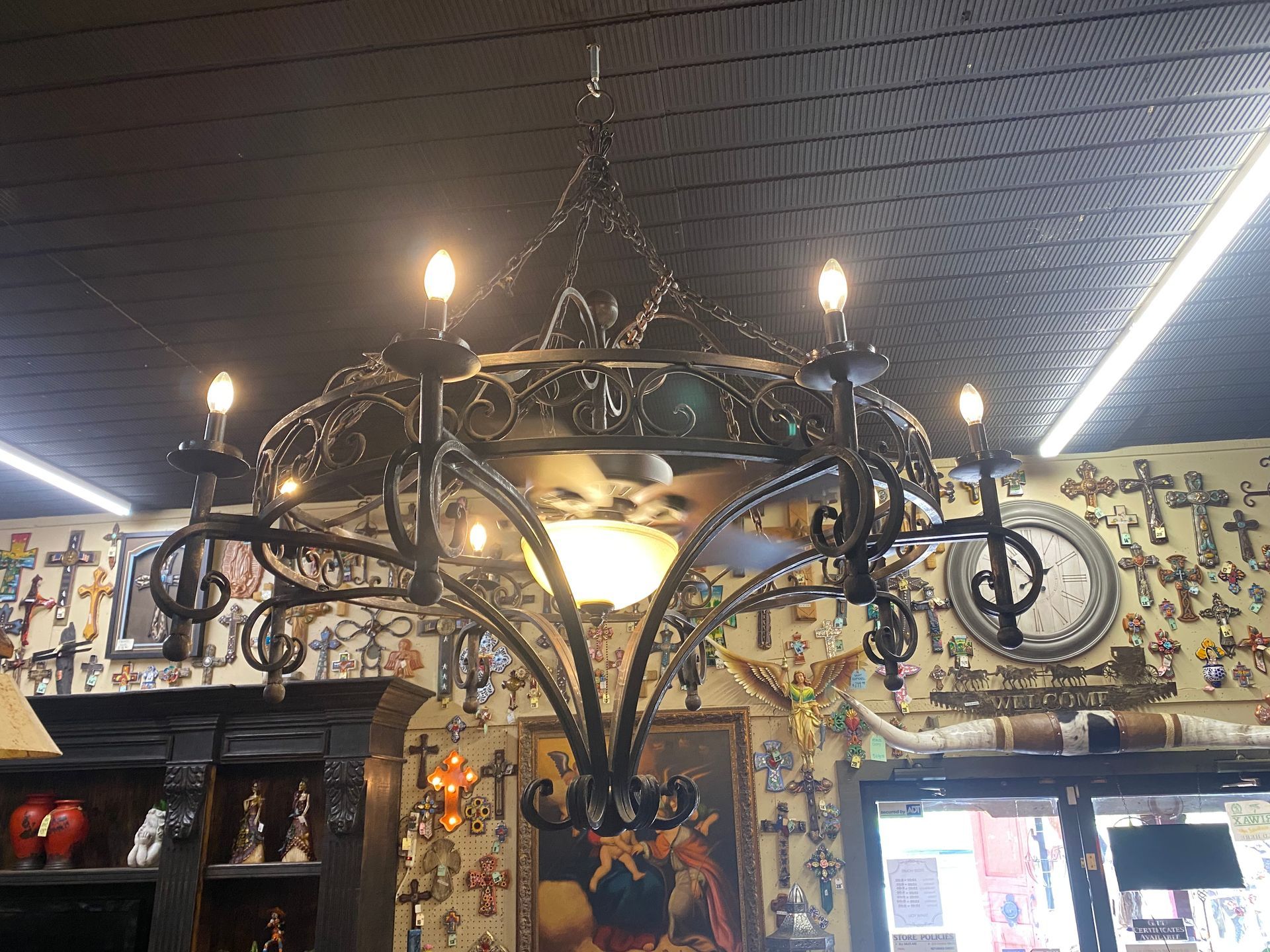 Ornate black wrought-iron chandelier with eight light bulbs, hanging in a store filled with religious decor.