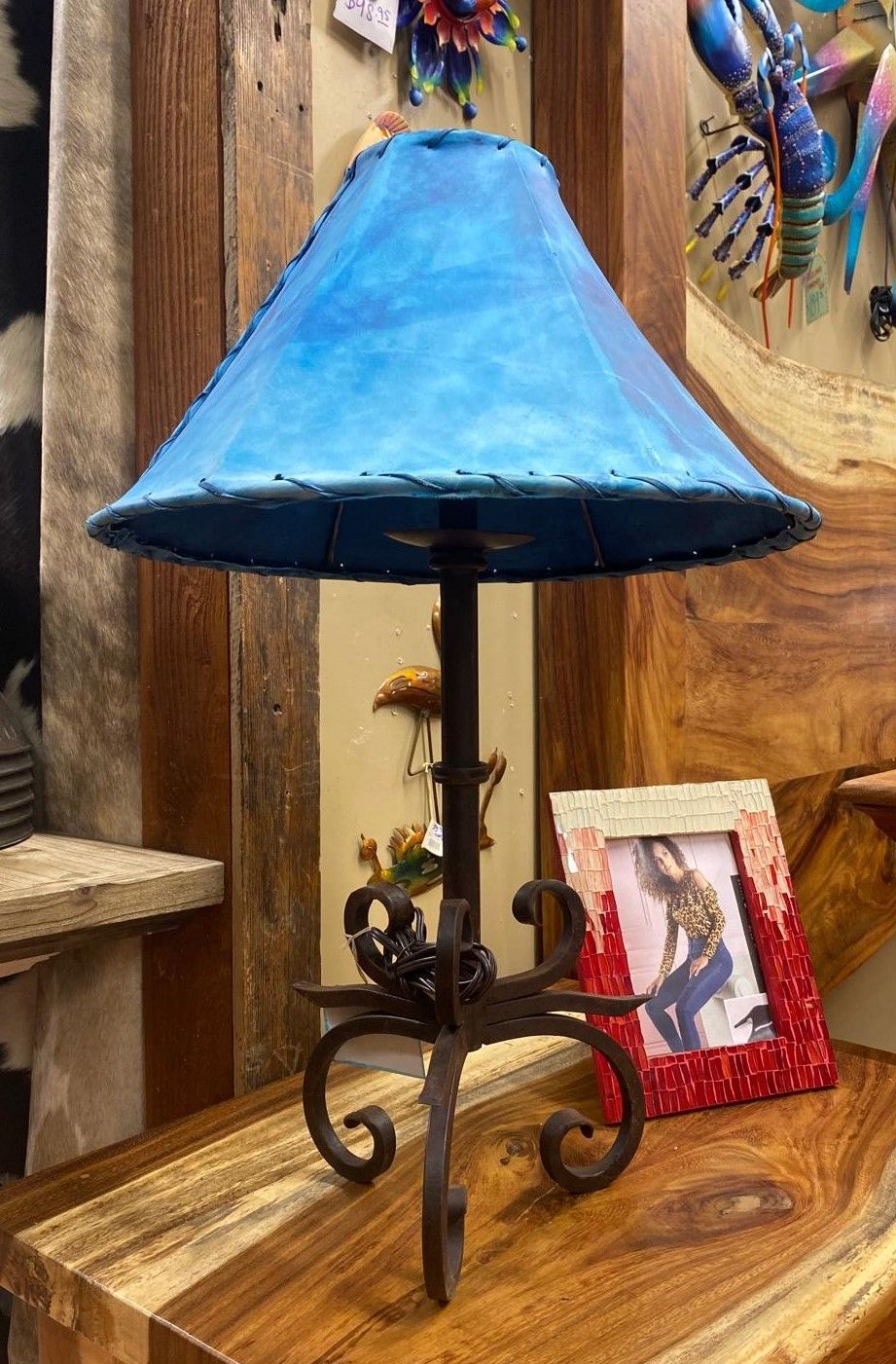 A rustic table lamp with a blue shade on a wooden surface. The base is wrought iron, and a framed photo is nearby.