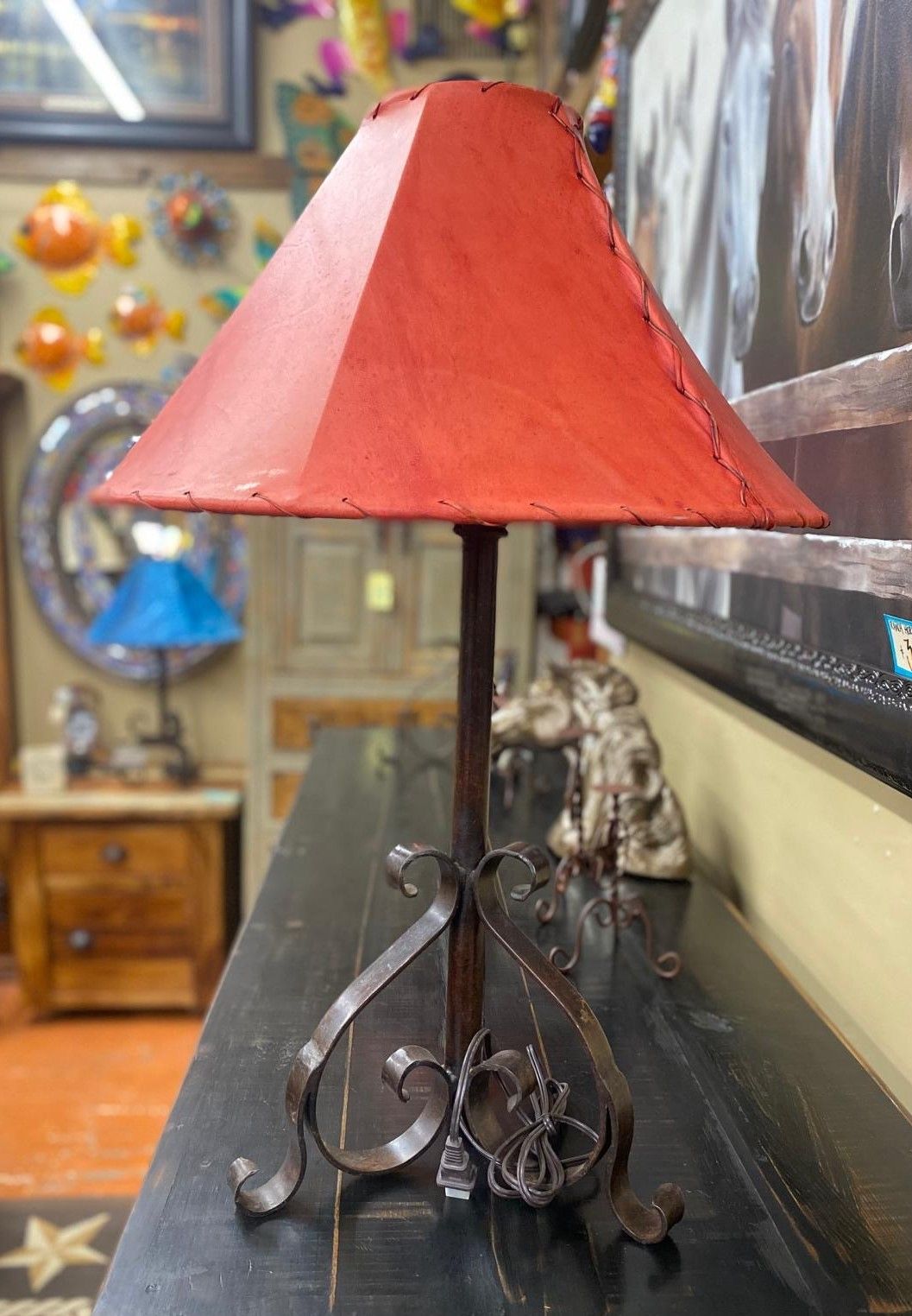A rustic red lampshade on a dark metal base, displayed on a black table in a shop setting with other decor visible in the background.