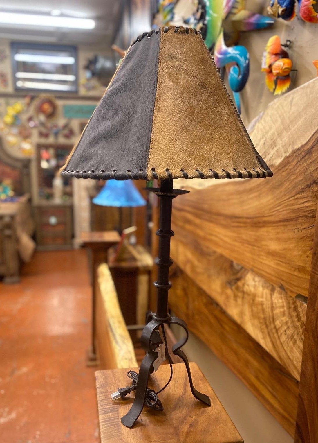 A rustic lamp with a two-toned leather shade sits on a wooden surface. It's in a shop with other furniture and art.
