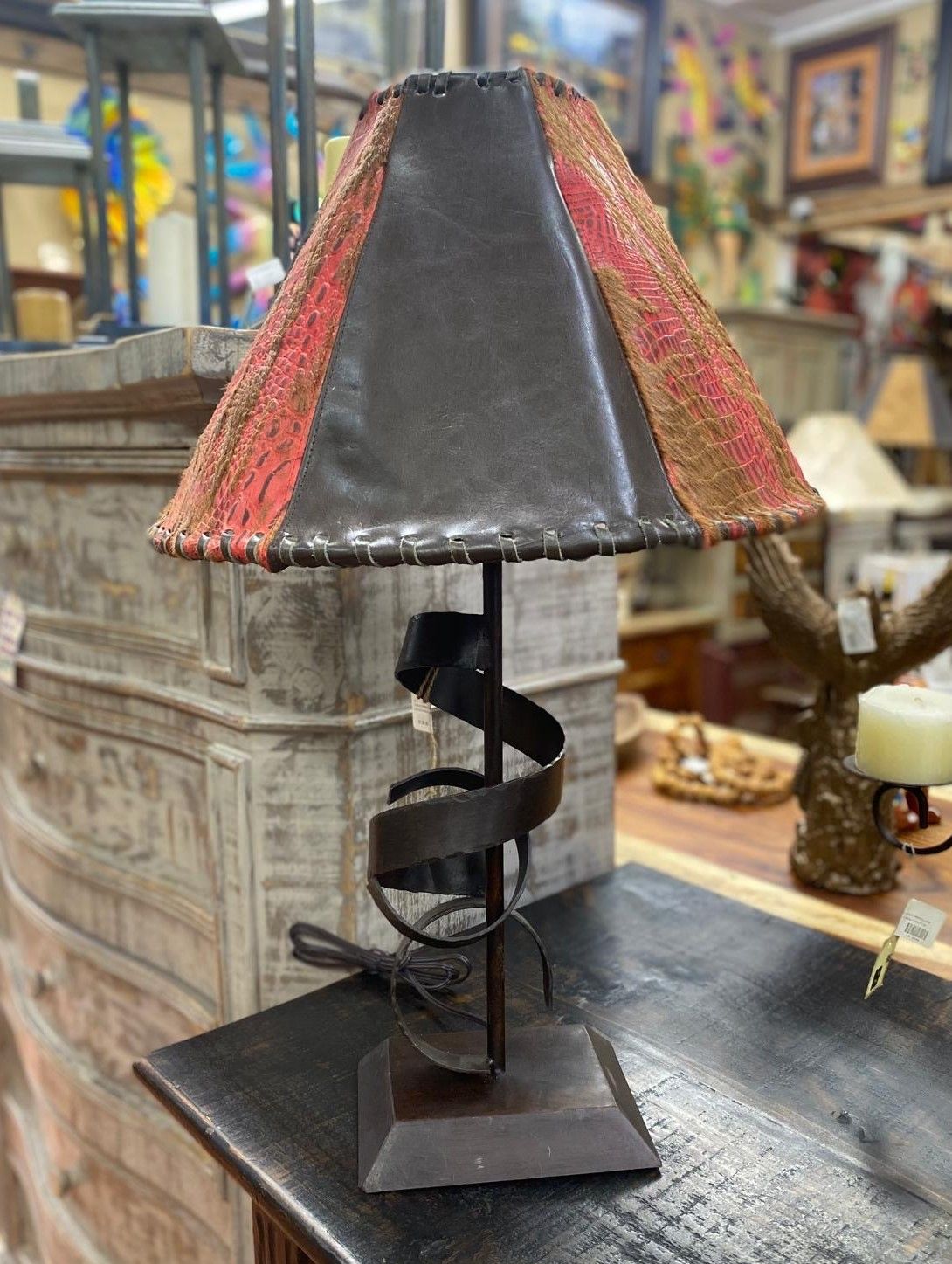 Table lamp with a brown, spiral-shaped metal base and a trapezoidal lampshade featuring leather and patterned fabric.