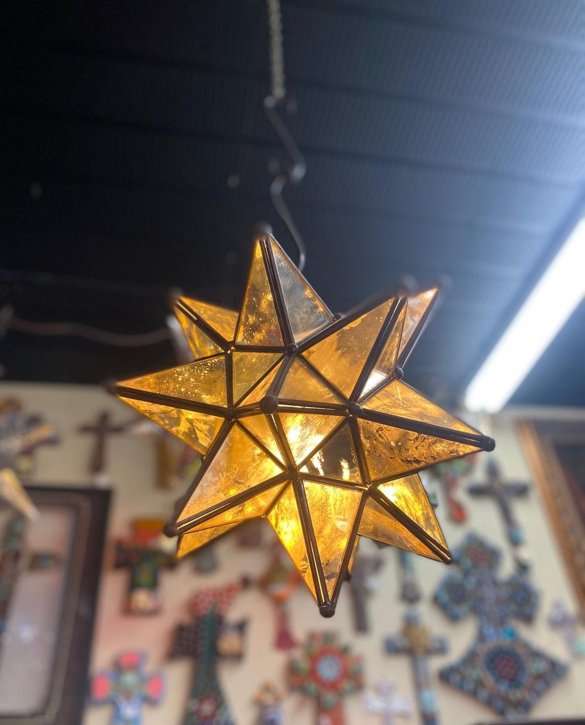 Star-shaped stained glass light fixture hangs indoors. The fixture has a warm yellow glow, with a background of decorative items.