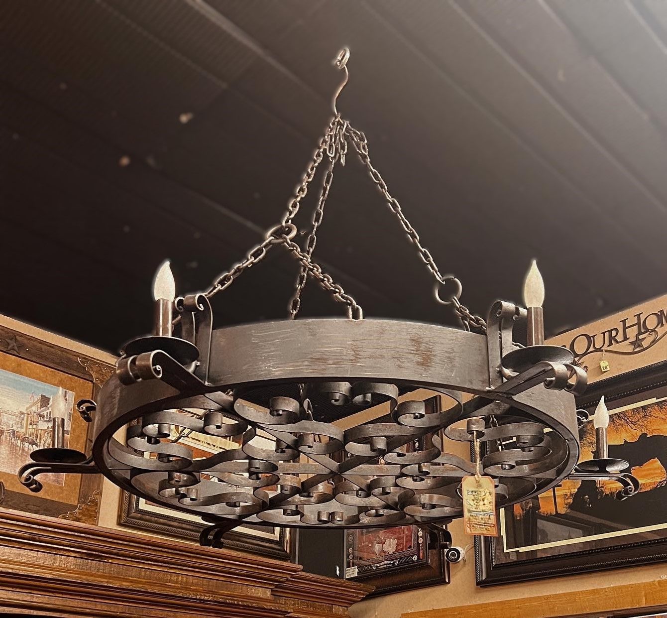 A wrought-iron chandelier with six faux candle lights hangs from the ceiling. The lamp is in a room with other decor on the walls.