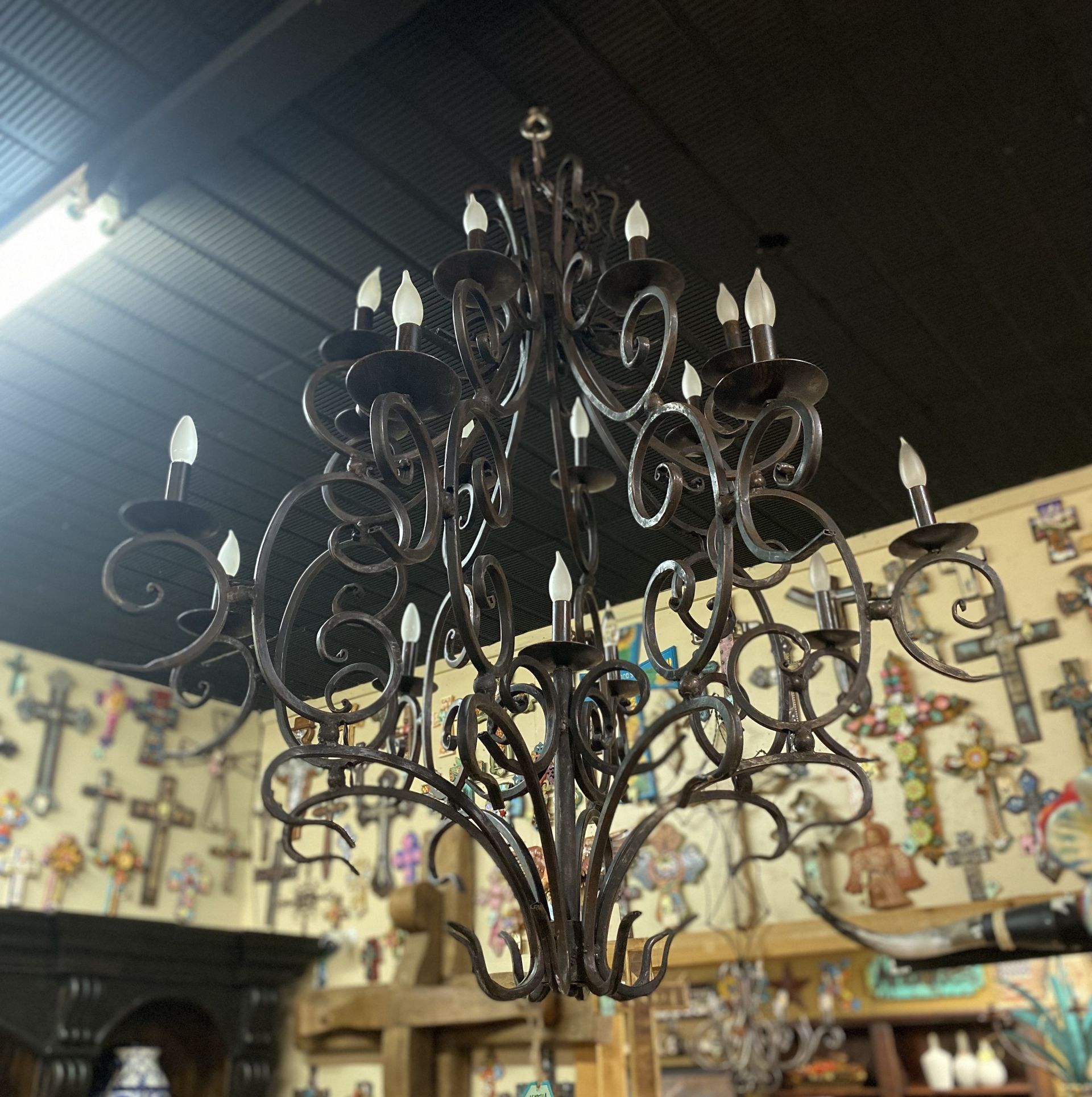 Ornate wrought iron chandelier hanging indoors, with many lights; store interior in background.