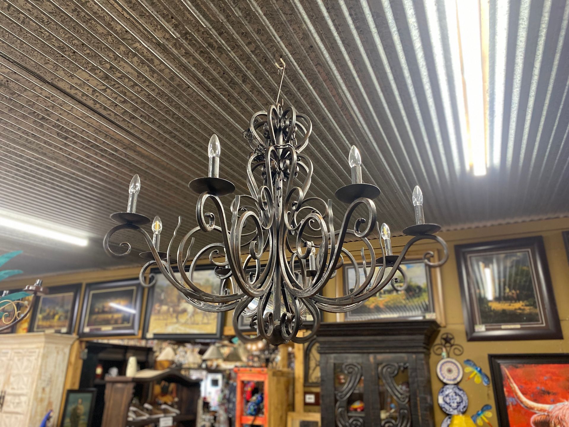 Ornate black wrought-iron chandelier hanging in a room with framed artwork and rustic furniture.