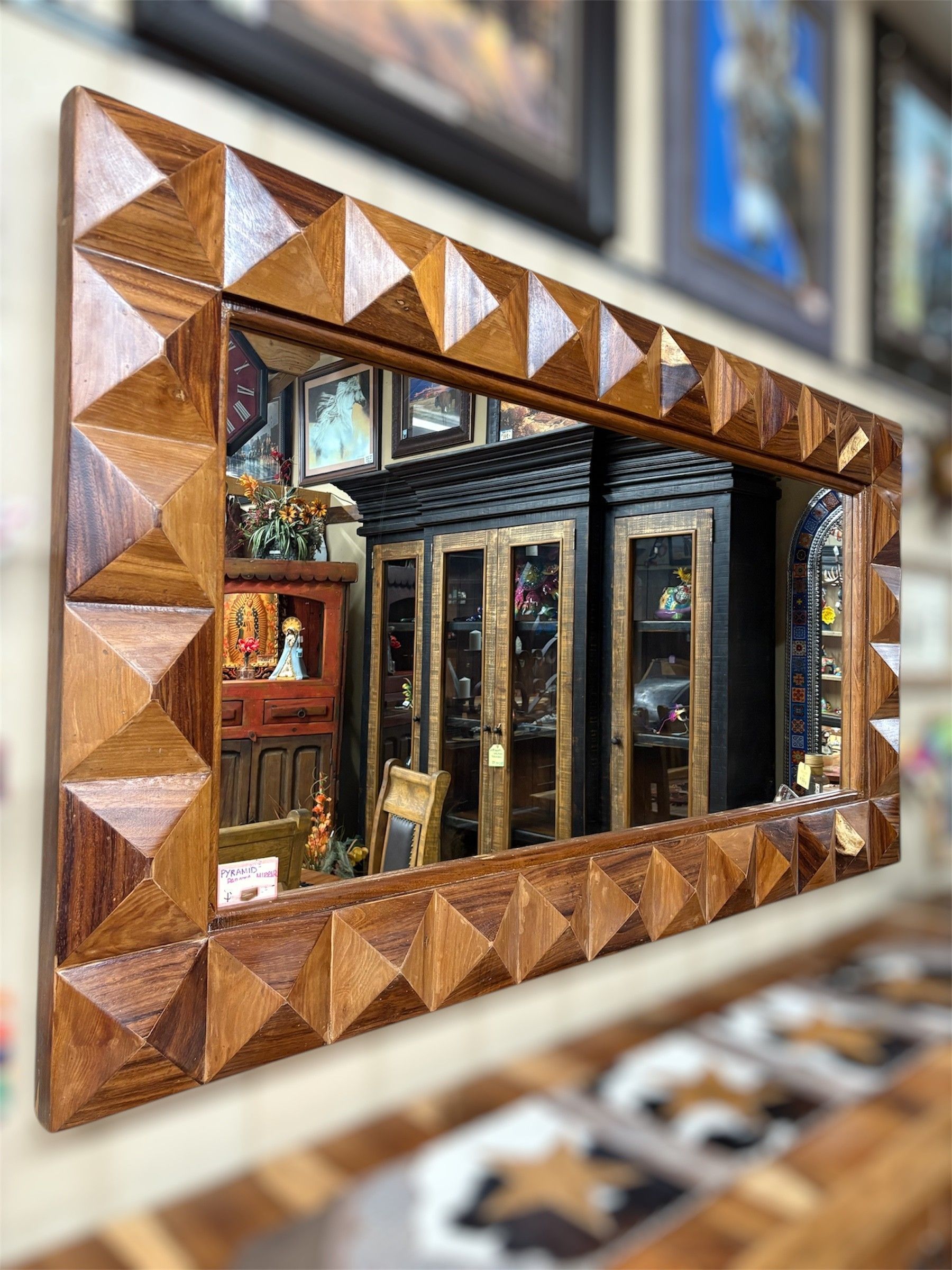 Rectangular mirror with a geometric, wood-carved frame hanging on a wall, reflecting a room with furniture and decorations.