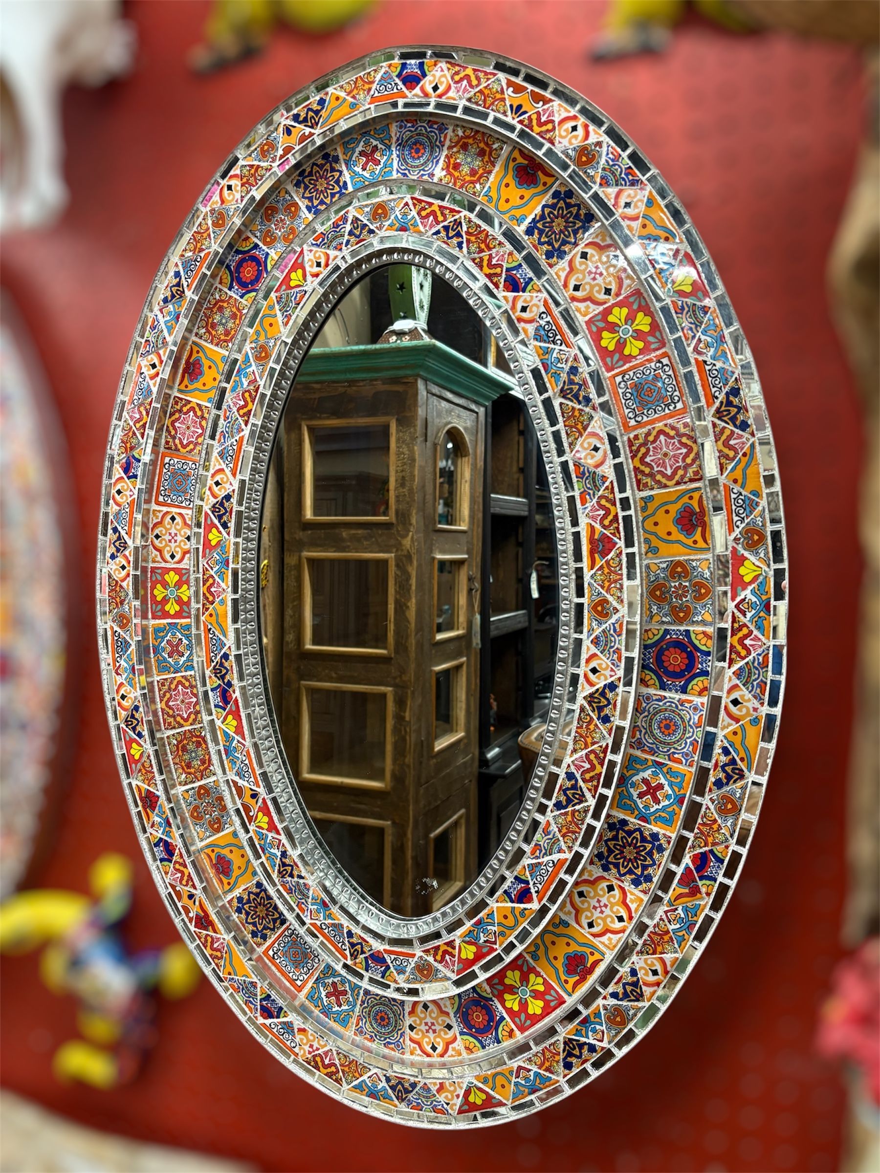 Oval mirror with a colorful mosaic tile frame, reflecting a dark wooden cabinet and room.
