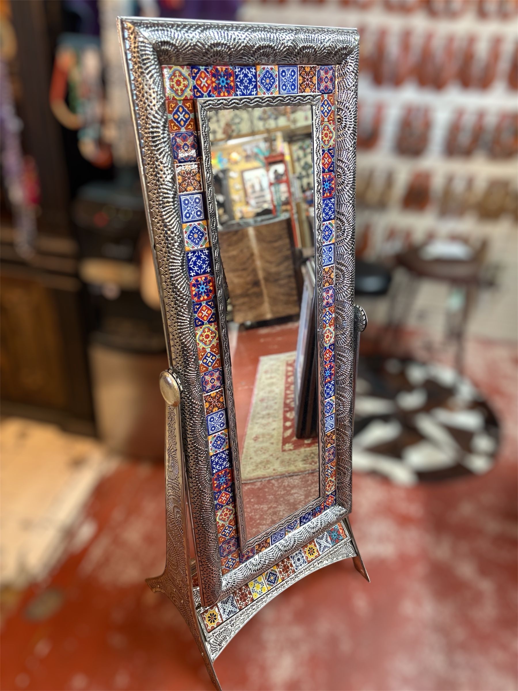 Ornate, tall standing mirror with a silver frame and colorful mosaic tile border, displayed on a red floor.