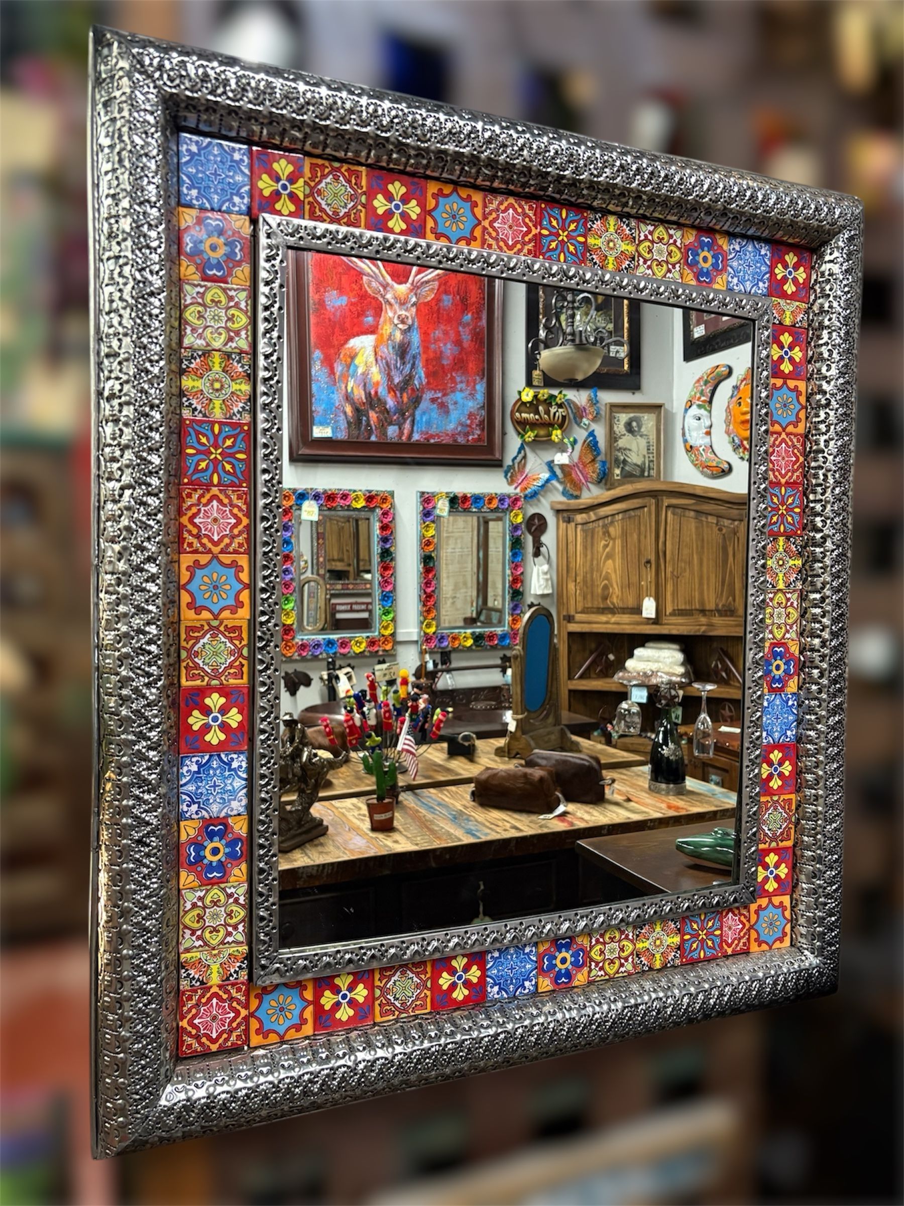 Square mirror with a decorative border of colorful tiles and a textured silver frame, reflecting a shop interior.