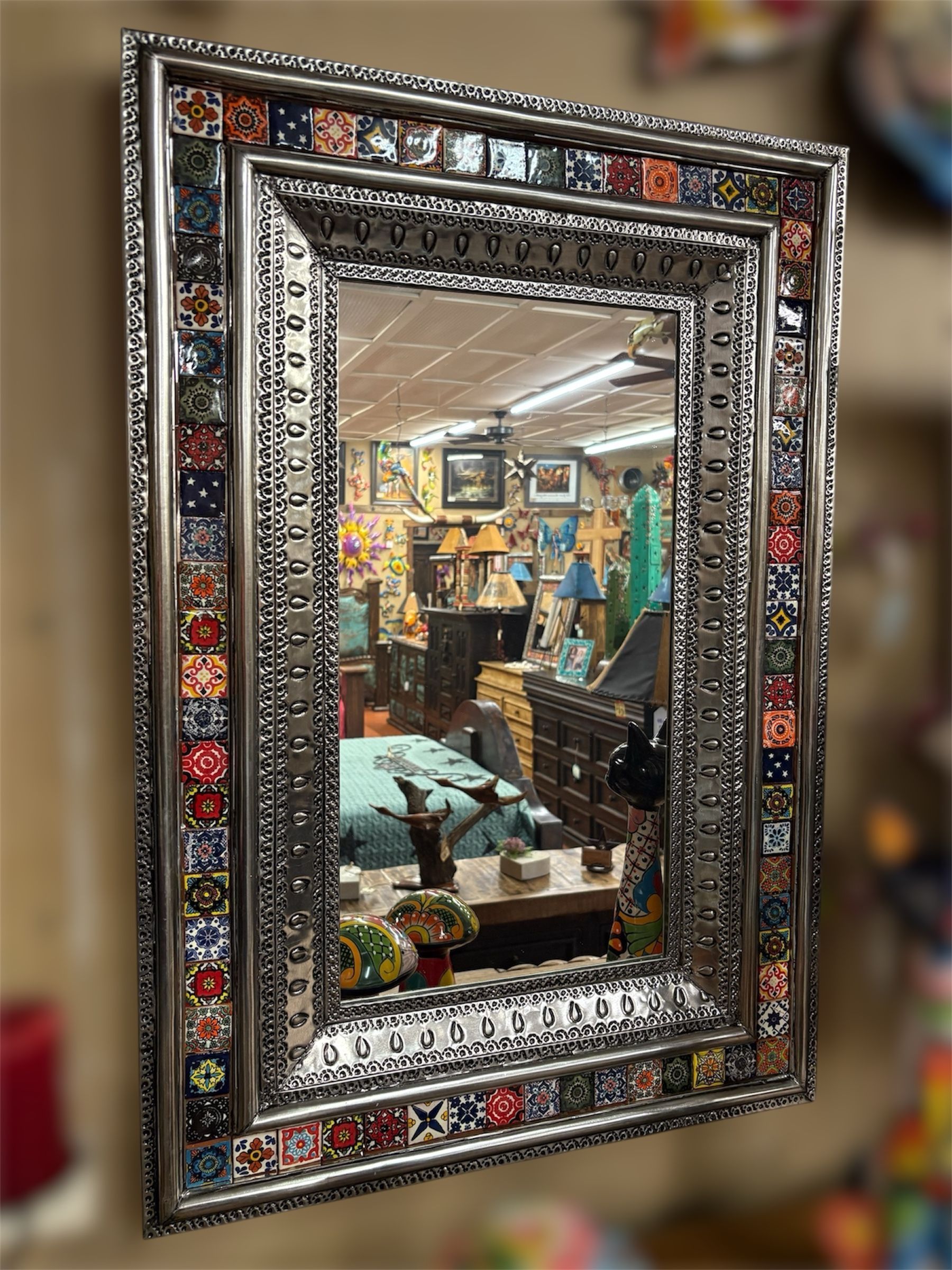 Ornate rectangular mirror with a decorative mosaic tile border, reflecting a cluttered shop interior.