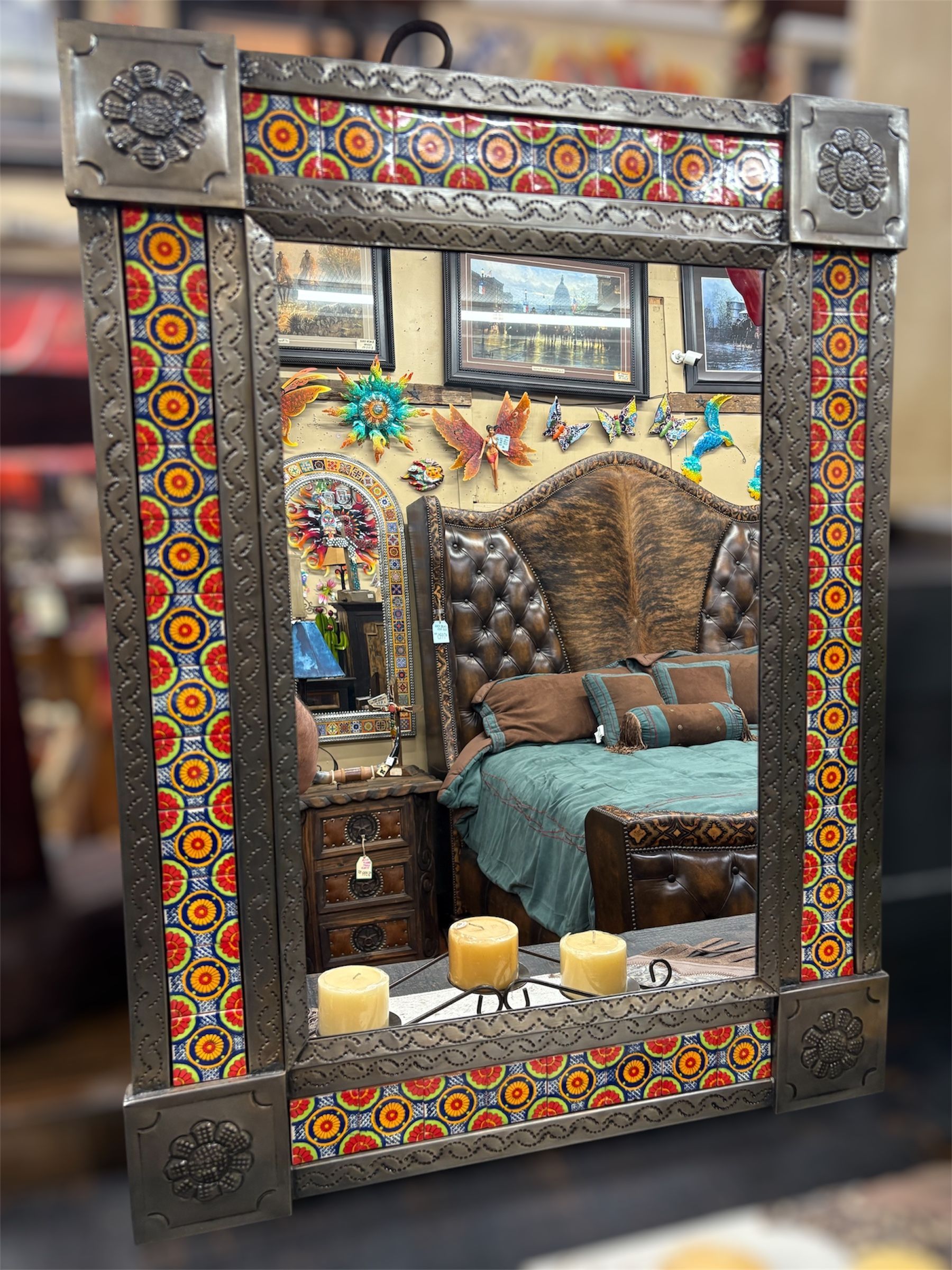 Ornate mirror with colorful mosaic tiles and metal trim, reflecting a bedroom with a tufted headboard and candles.