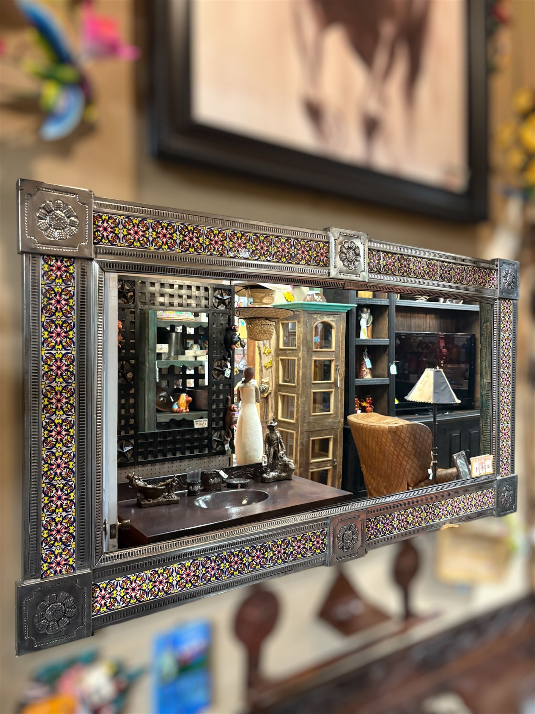 Ornate mirror reflecting a room scene: a figure stands near a door, with shelves and a desk visible. The frame is textured with metallic and colorful accents.