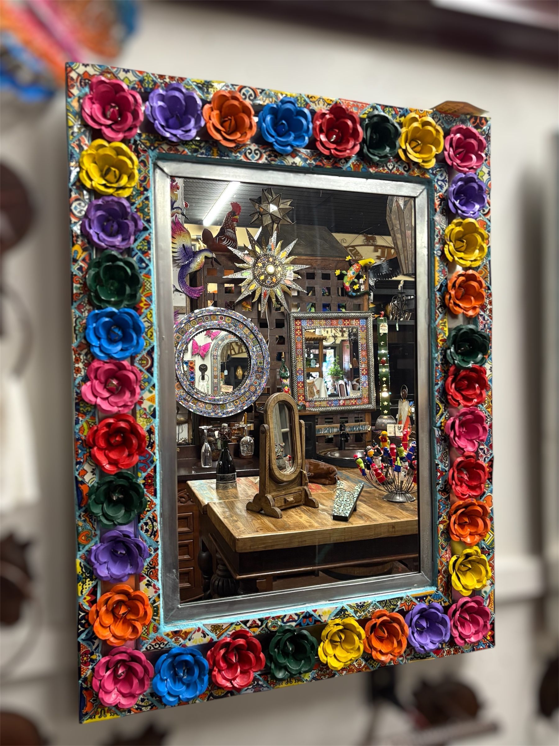A colorful mirror with a frame decorated with rows of paper flowers. The reflection shows an antique store interior.