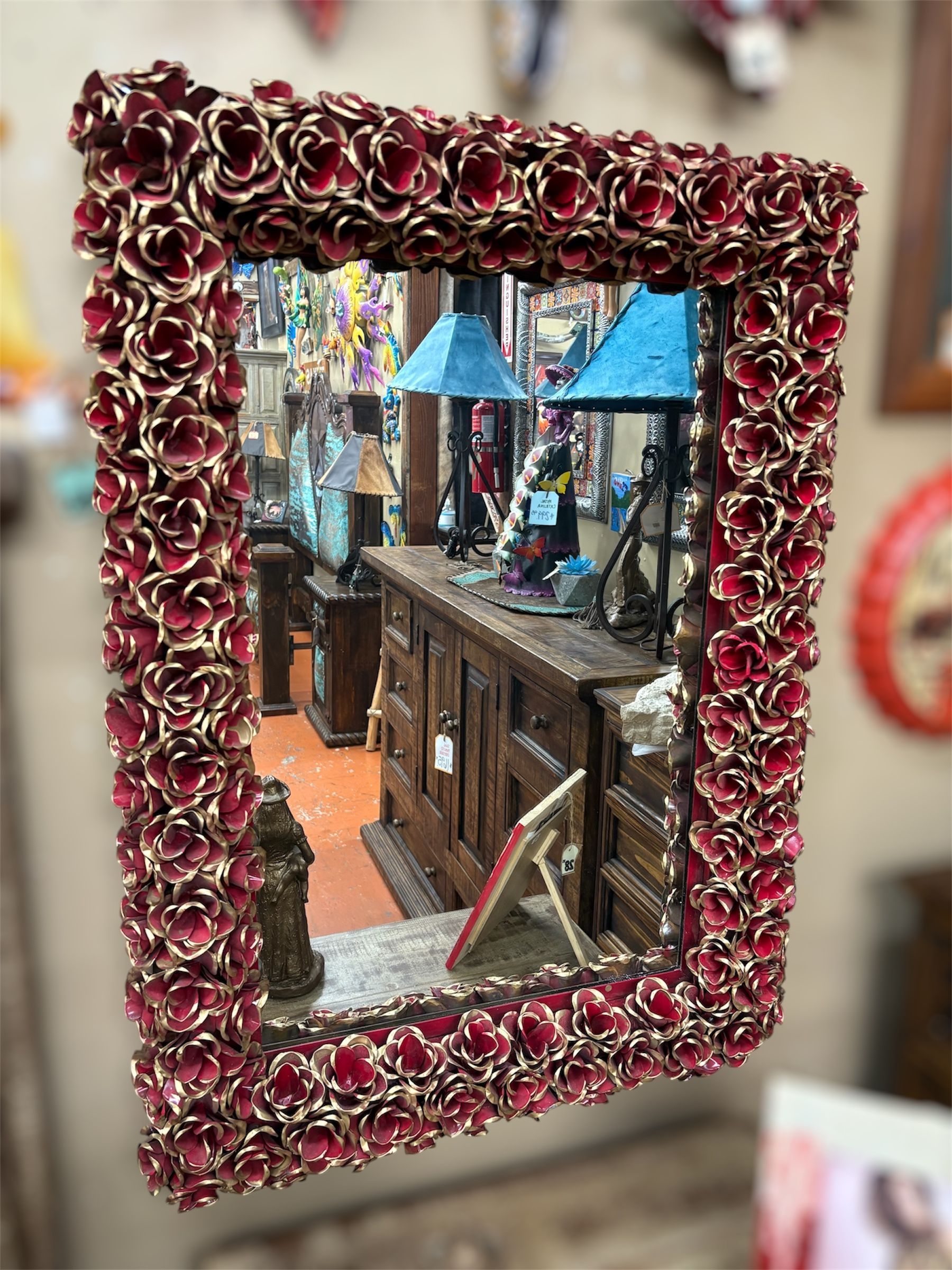A rectangular mirror with a frame of red and gold flowers reflects a cluttered shop interior.