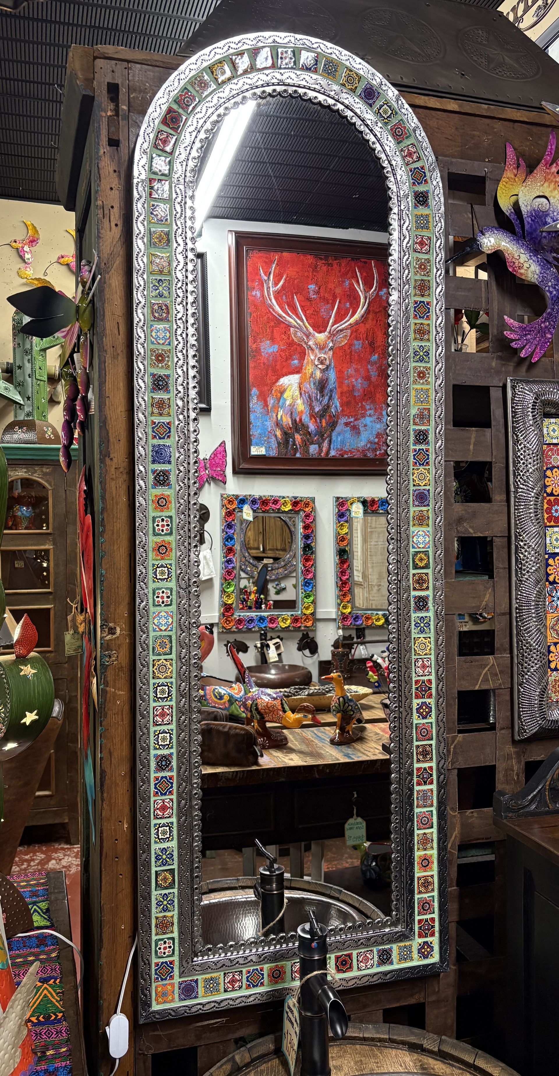 Tall, arched mirror with a mosaic tile border in a shop setting. Reflected in the mirror are colorful art and objects.
