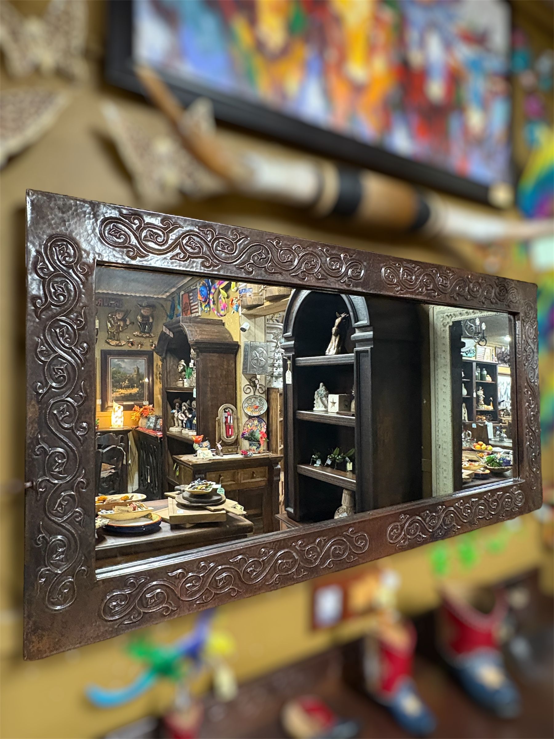 Ornate rectangular mirror reflecting a room filled with antiques and decorative items. The frame is dark brown with detailed carvings.