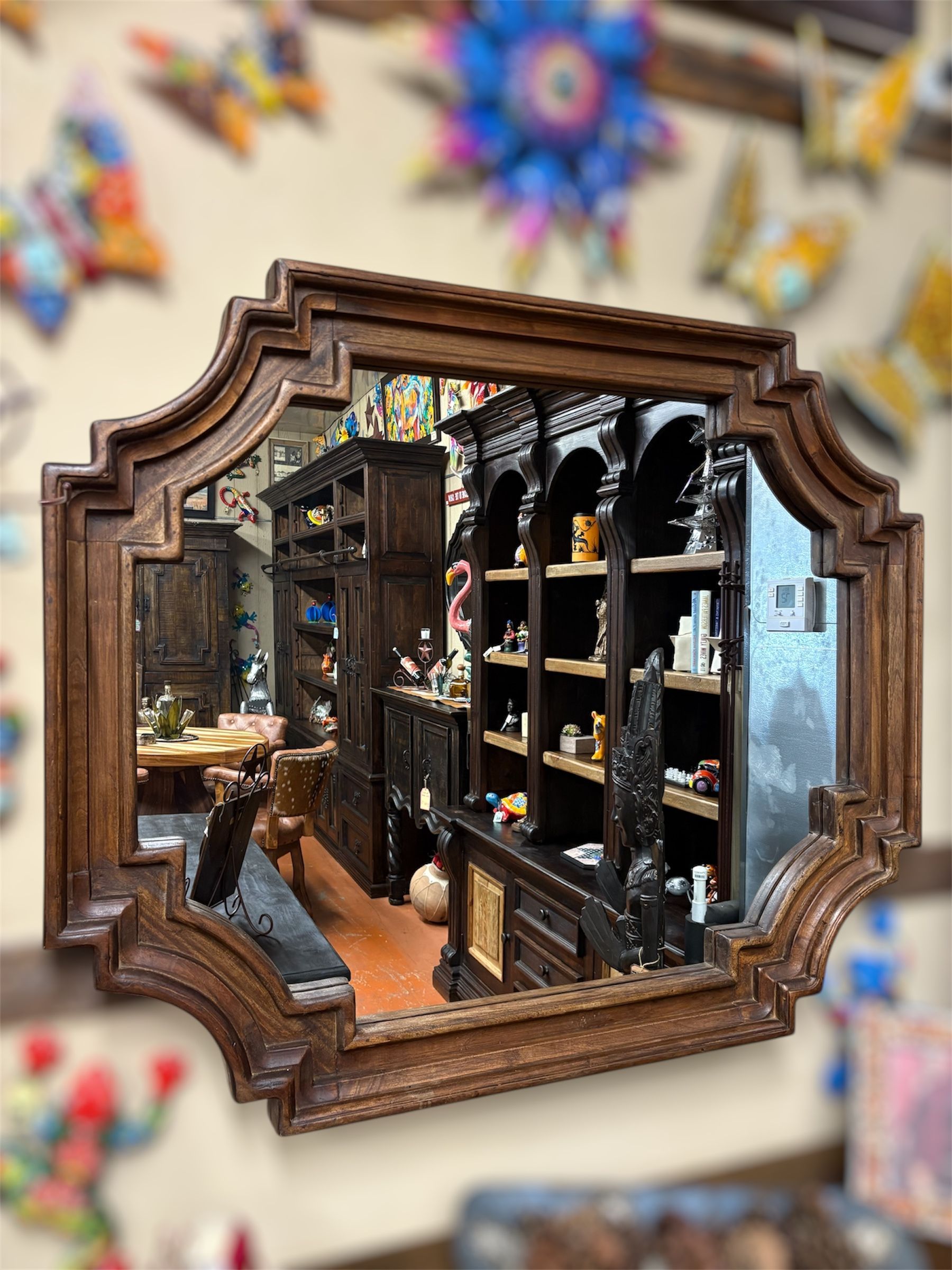 Wooden framed mirror reflecting a room filled with shelves and objects; colorful decorations are in the blurred background.