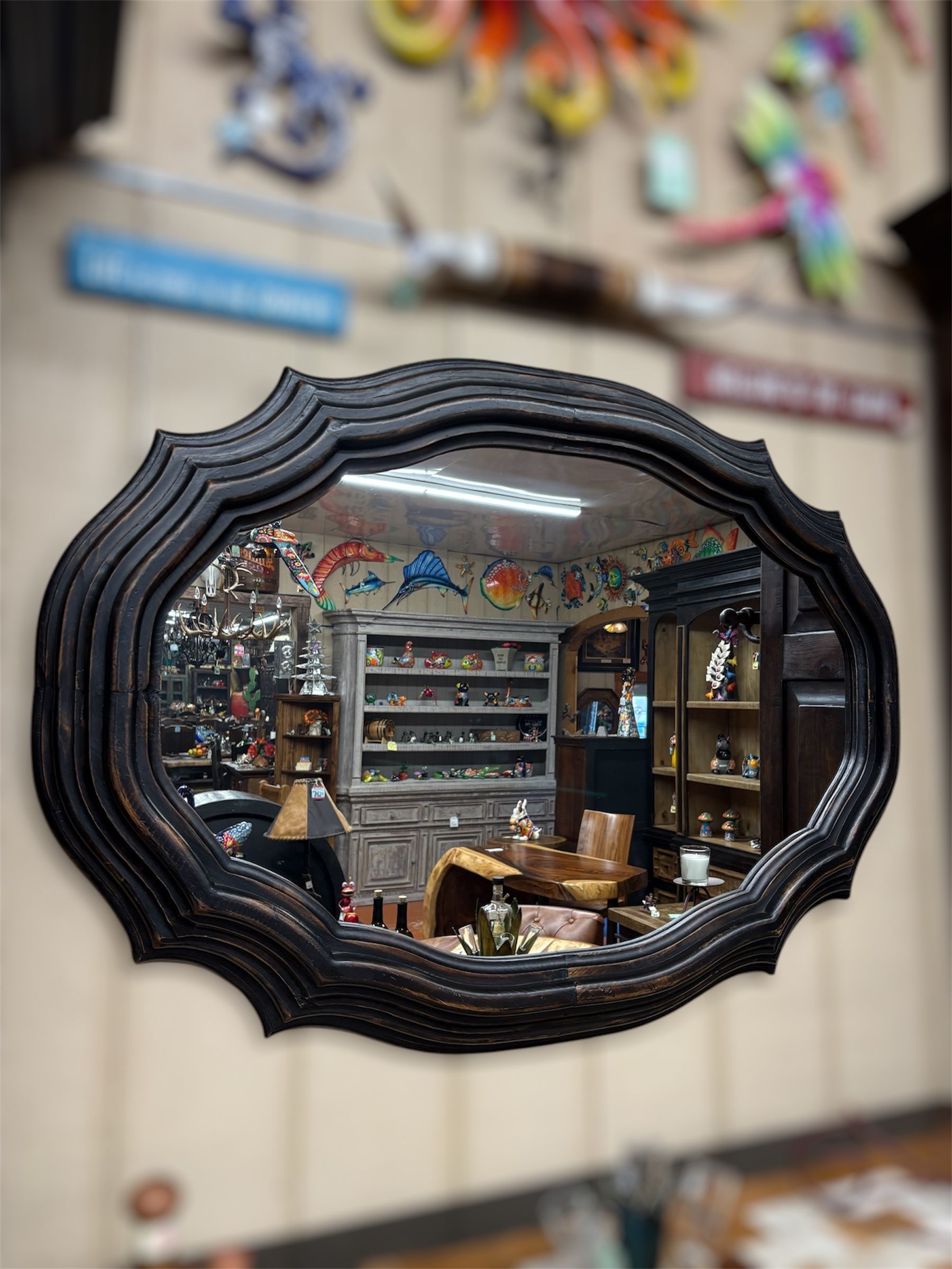 Black ornate mirror reflecting the interior of a shop, with shelves, figurines, and artwork.