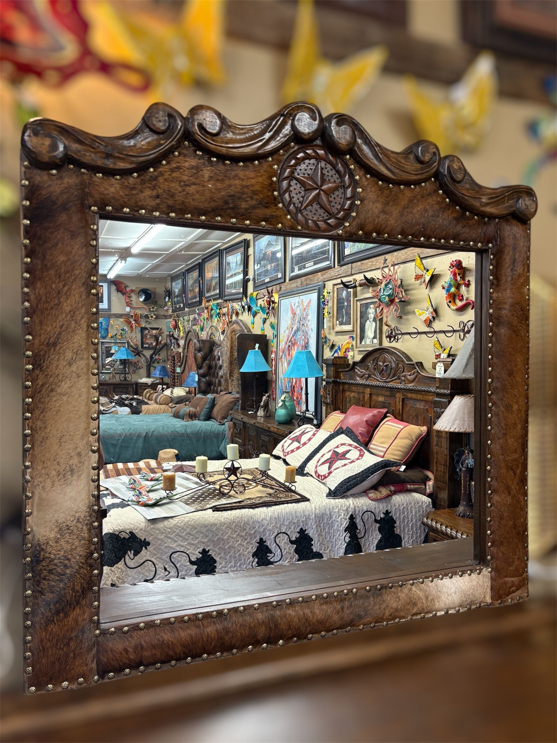 Large decorative mirror with a leather-like frame, reflecting a rustic furniture store's interior.
