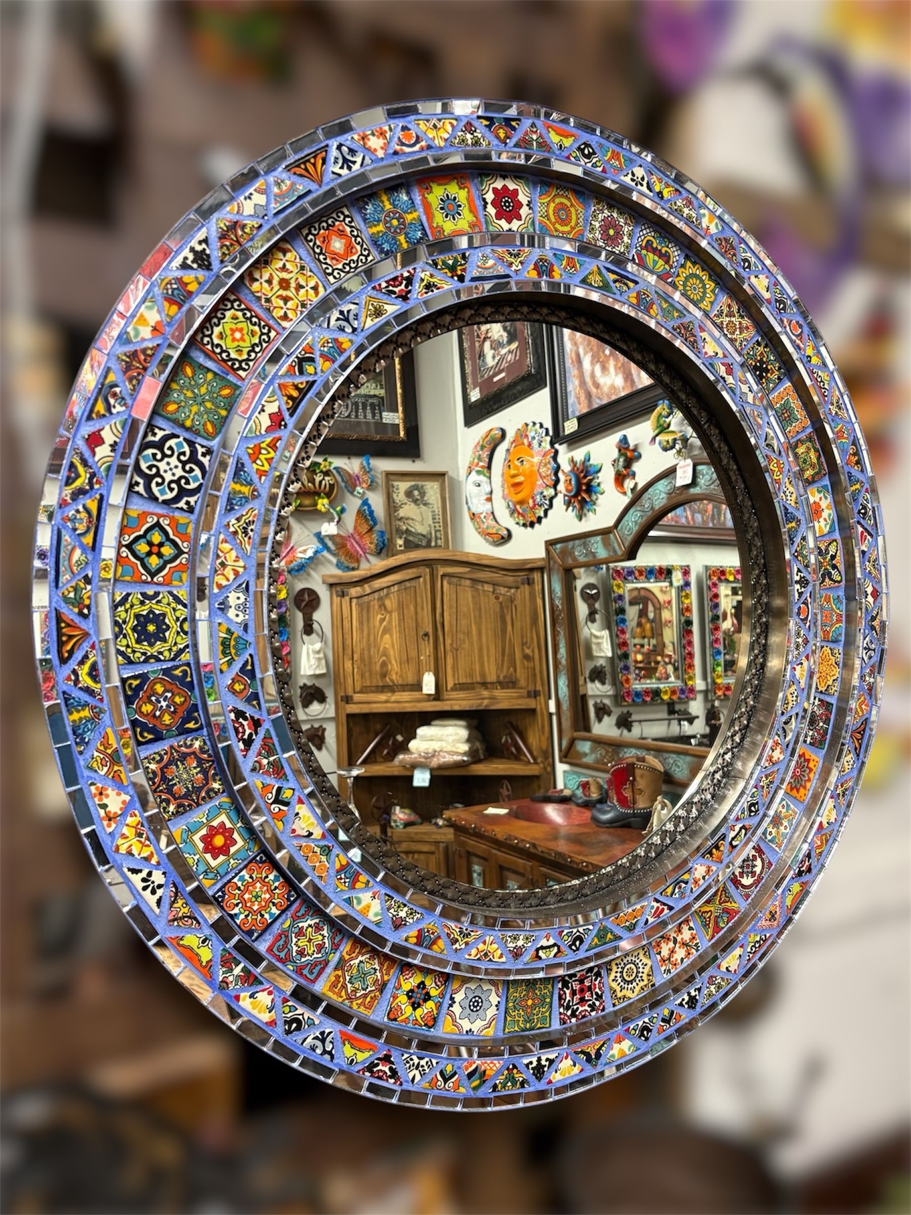 Round mirror with mosaic tile border.  Reflects a room filled with artwork, furniture, and a wooden cabinet.