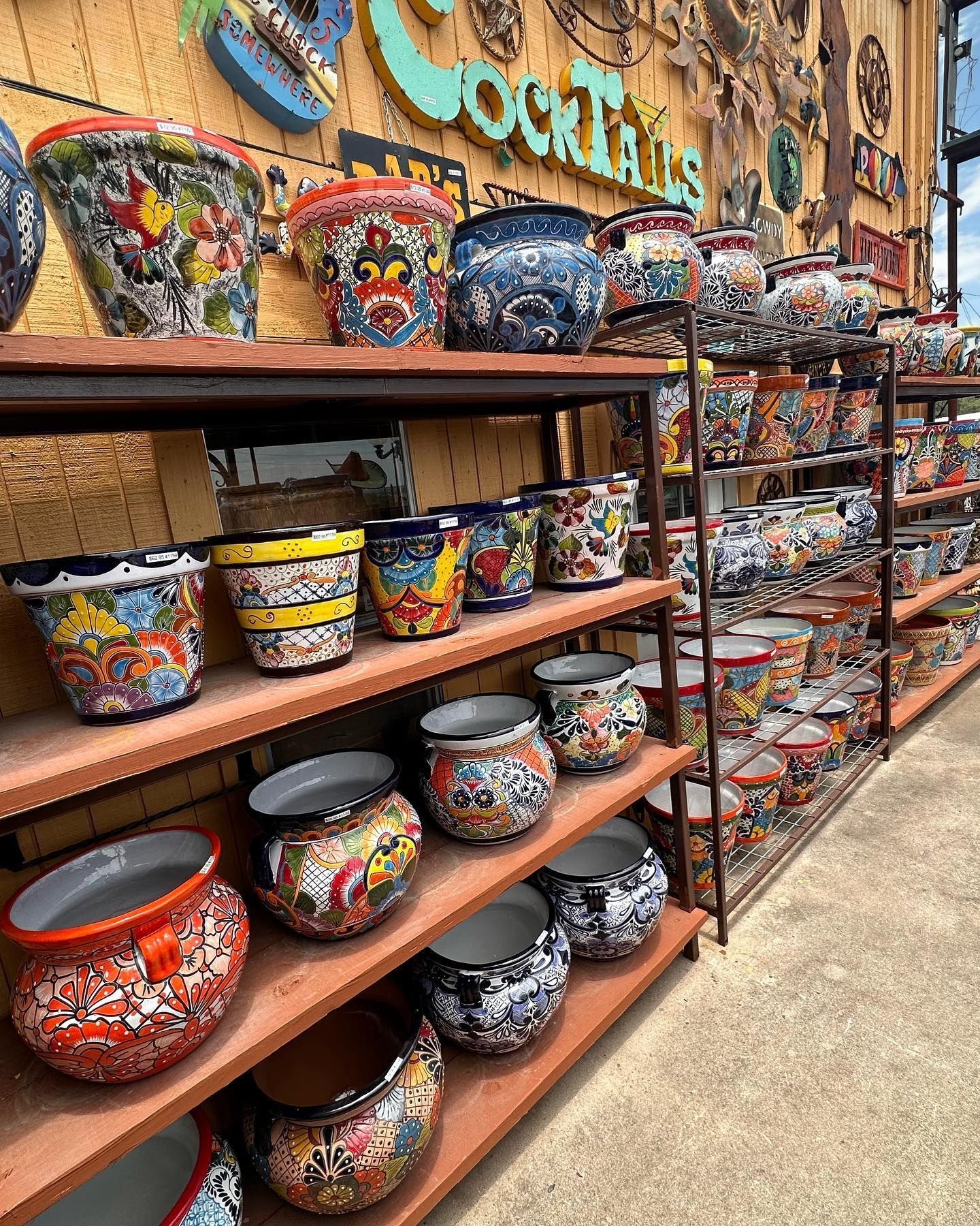 Shelves displaying colorful, patterned ceramic pots at an outdoor shop; the backdrop features a wooden wall and Cocktails sign.
