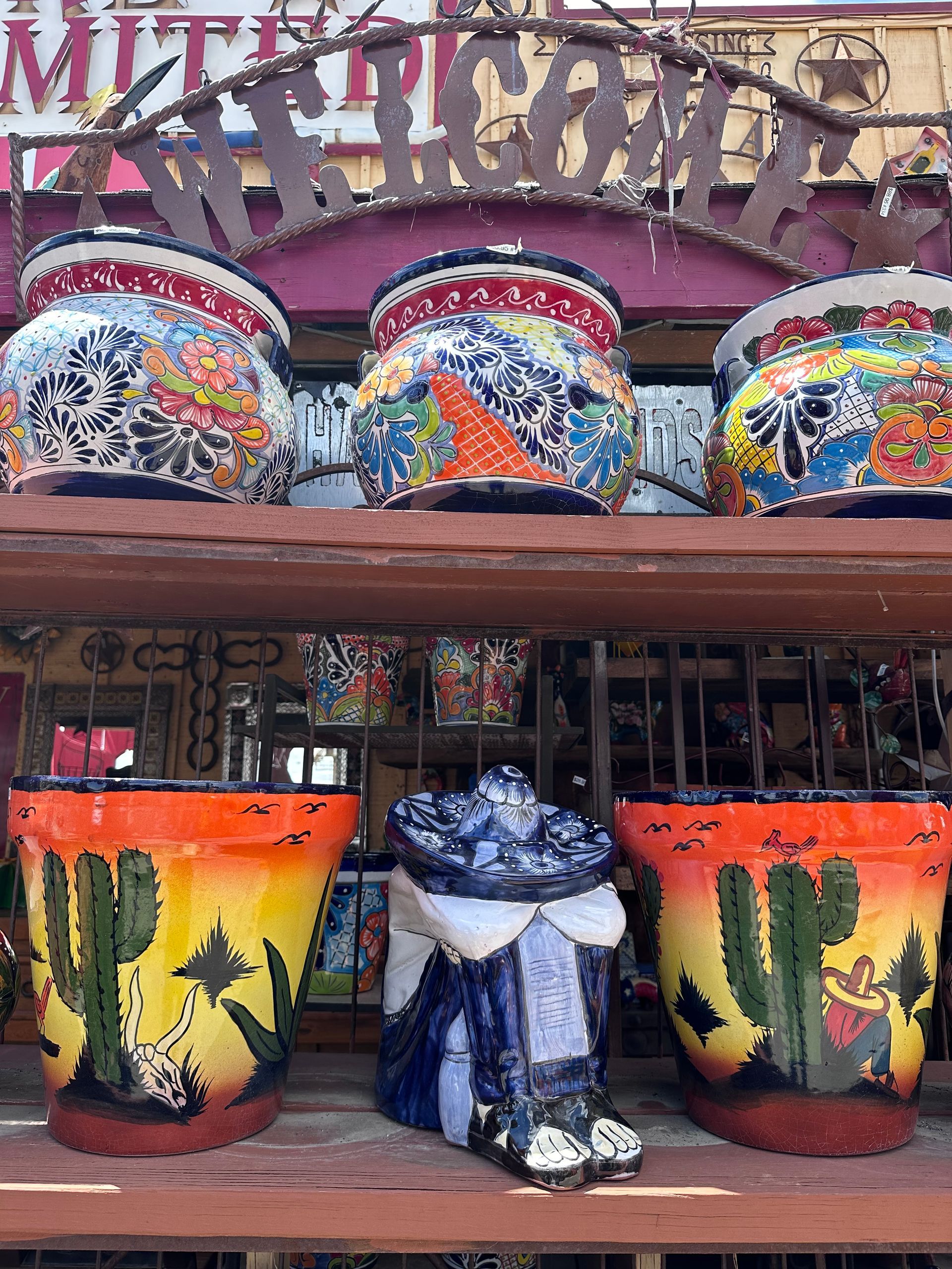 Shelves displaying colorful, hand-painted Mexican pottery. The top shelf has three large pots, while the bottom has three pots and a boot.