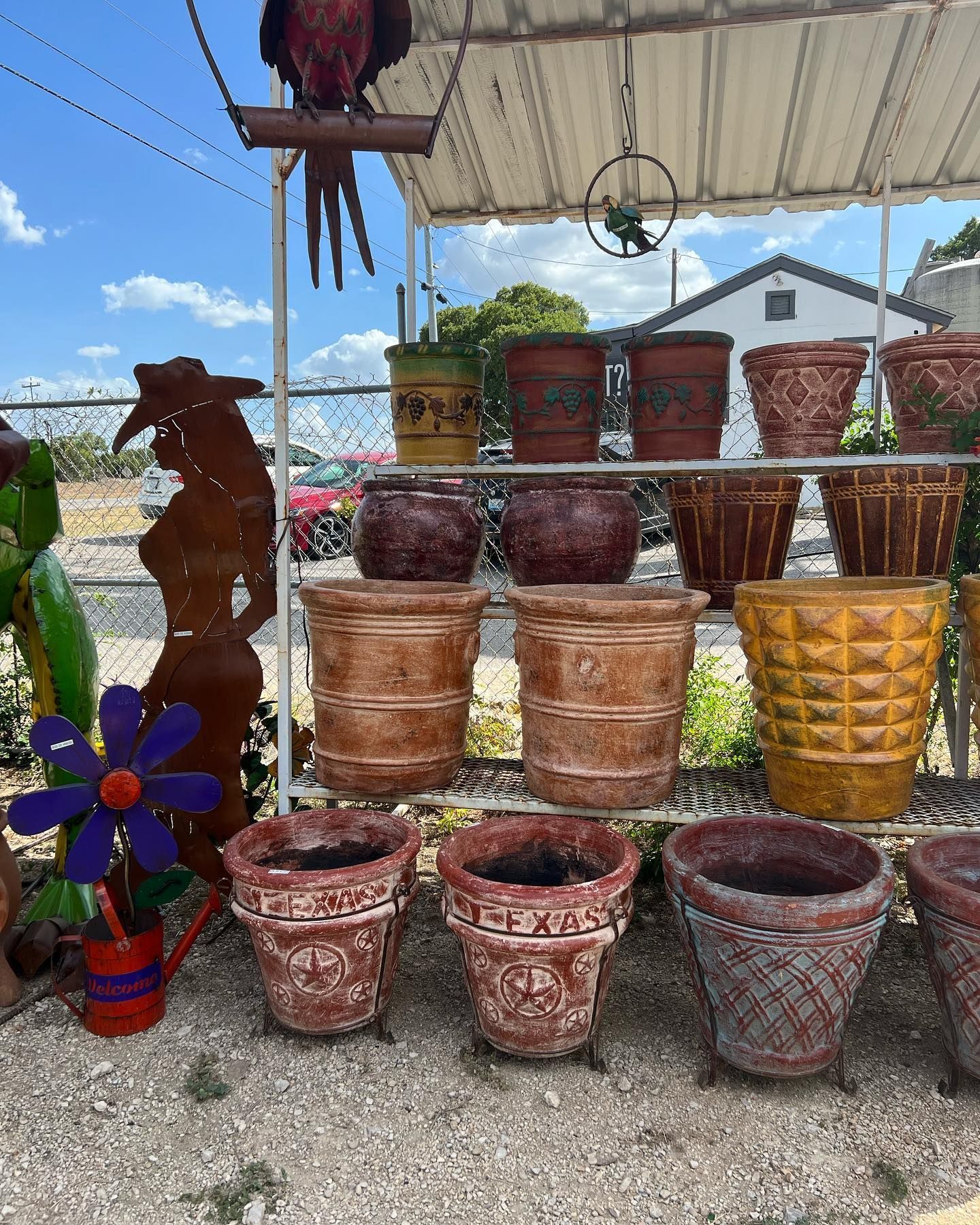 Rows of various terracotta flower pots on display at an outdoor store, alongside metal art and a parrot statue.