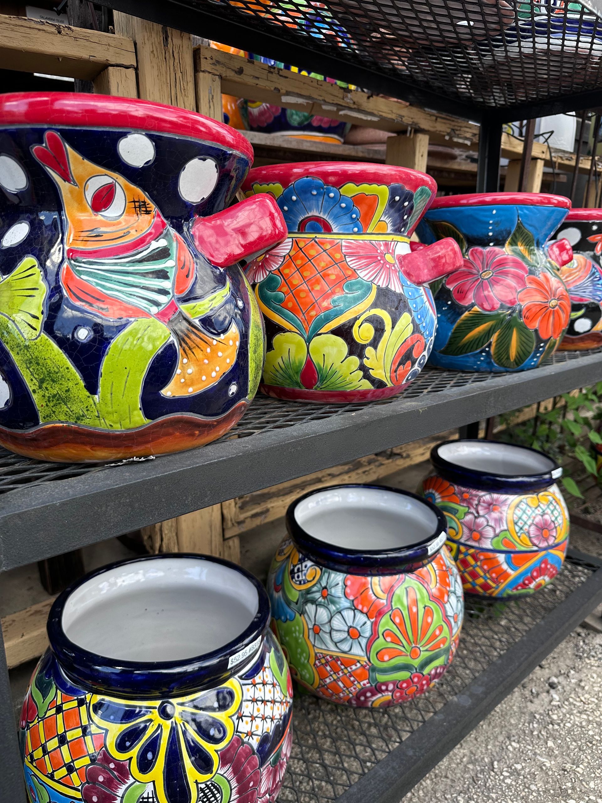 Colorful, hand-painted Mexican pottery for sale, displayed on metal shelves. The pots feature vibrant floral and animal designs.