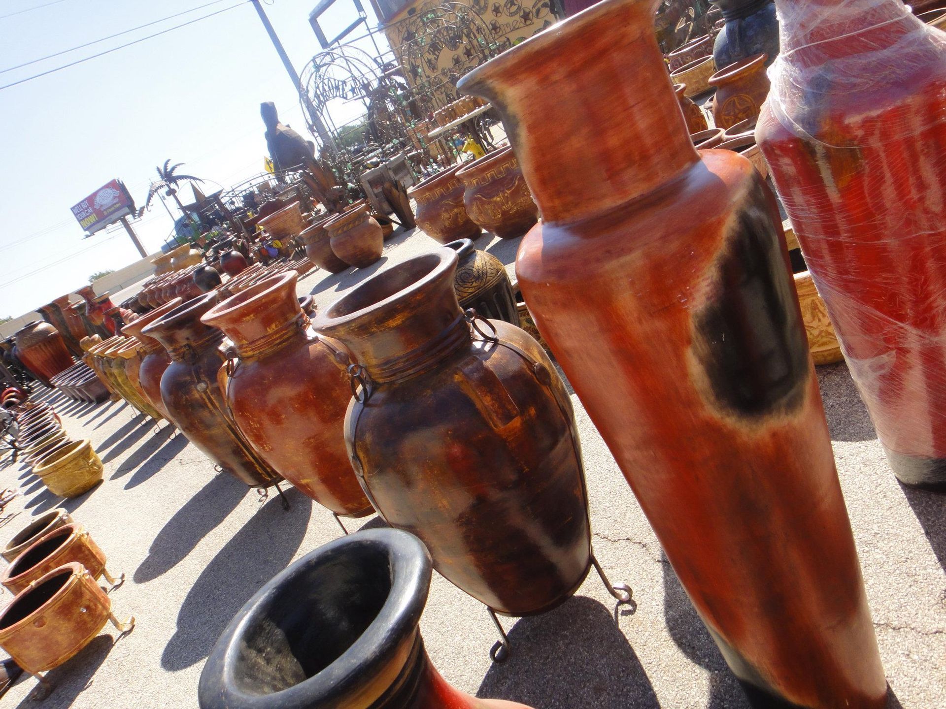 Rows of large, glazed ceramic pots in shades of brown and orange, displayed outdoors. Sunlight casts shadows.