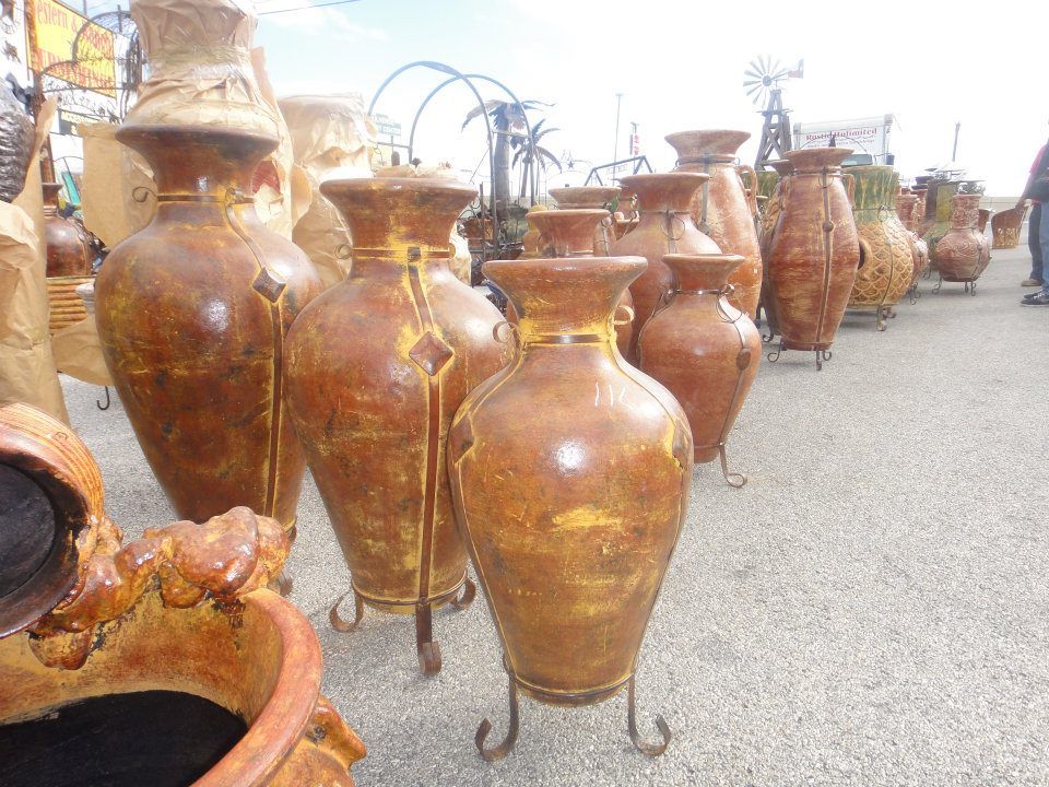 Rows of large, weathered brown pottery vases on metal stands, displayed outdoors in what appears to be a marketplace.