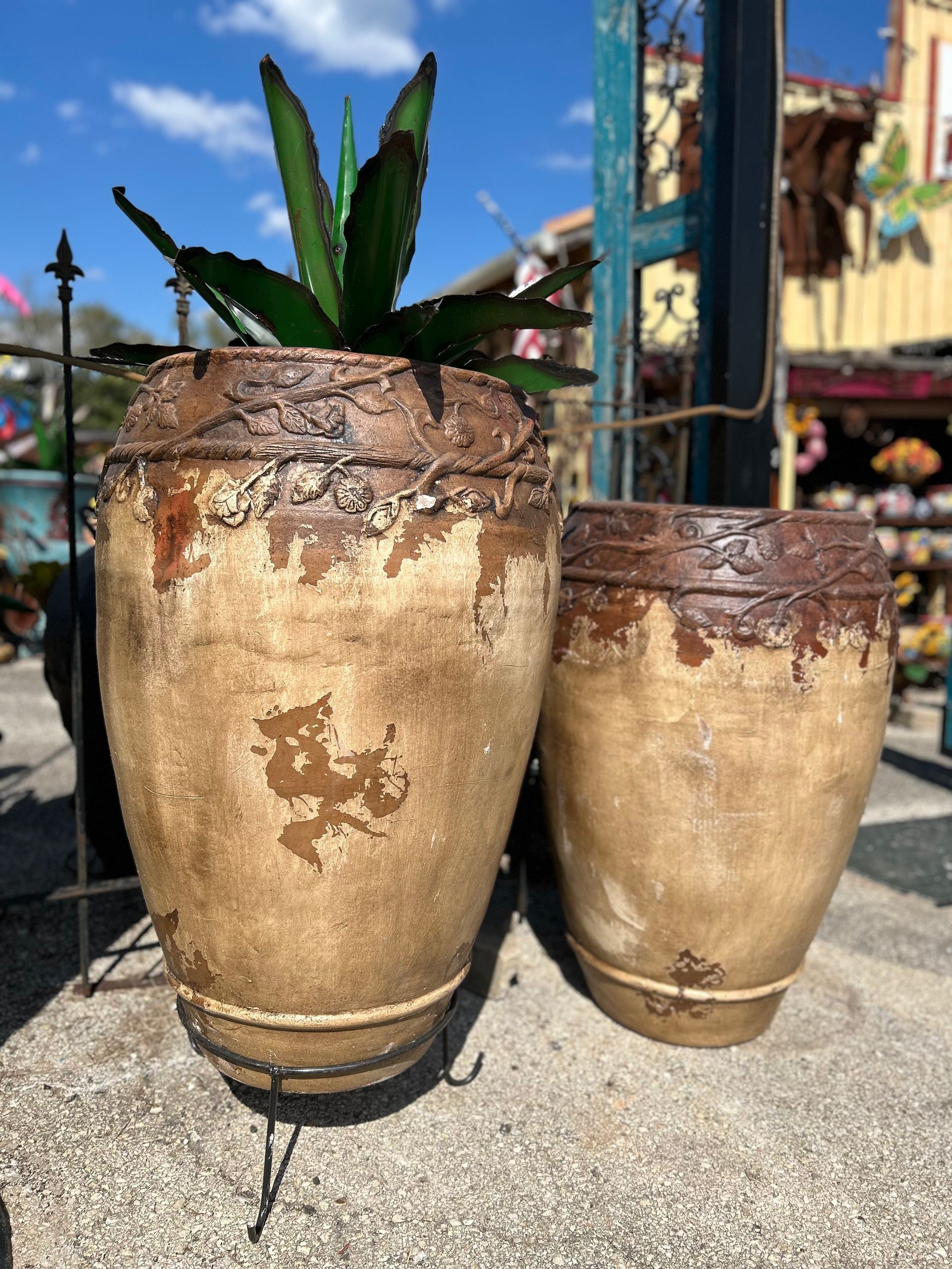 Two aged, tan-colored planters with dark brown rims and detailing. One contains a green plant. They sit outdoors.