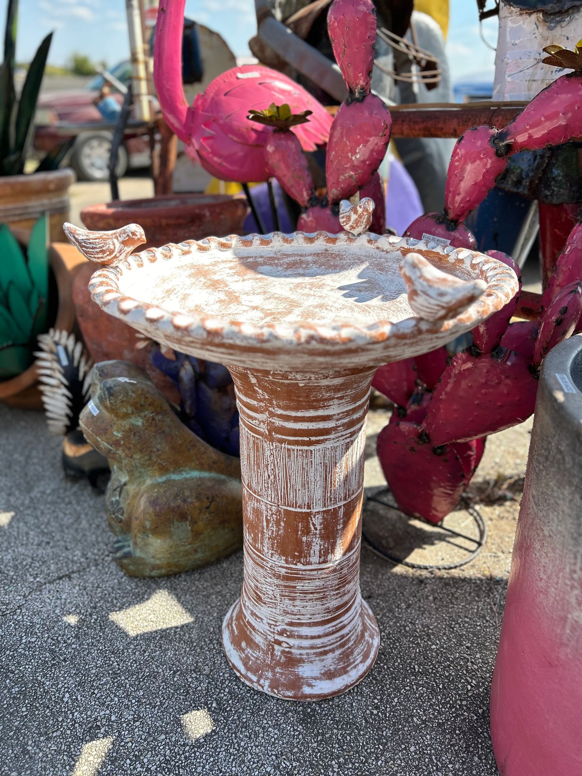 A terracotta birdbath with white accents stands outside among garden decor. A pink flamingo statue is visible in the background.