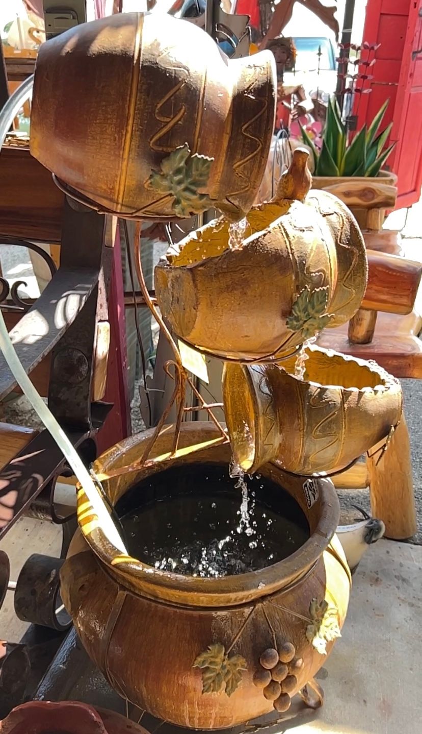 A cascading water fountain with four stacked, barrel-shaped containers. Water flows from the top to a large basin below, with decorative leaf and grape details.