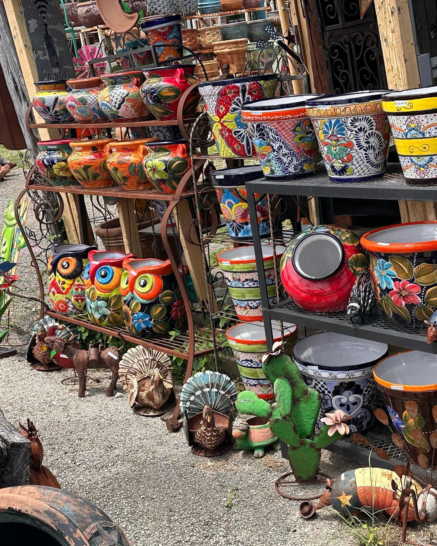 Shelves displaying colorful ceramic pots and sculptures outdoors, including owls, cacti, and planters with intricate designs.