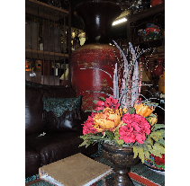 A dark red ornate vase sits near a couch with colorful flowers and a book on the coffee table.