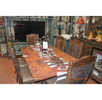 Rustic dining room set with a large wooden table, leather chairs, and a southwest-style runner. The setting includes a TV, decorative shelves, and artwork.