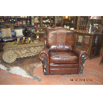 Brown leather recliner chair with decorative side panels in a rustic-themed store. A carved coffee table and cowhide rug are nearby.