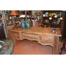 Rustic wooden desk with drawers and a curved front, inside a room with shelves and decor.