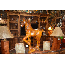 A large, wooden horse sculpture stands in a rustic-looking room, with wood furniture, lamps, and shelves filled with items.