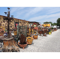 Outdoor antique market, showcasing metal sculptures, planters, and various items in front of a weathered building, under a sunny sky.
