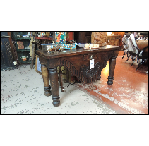 Wooden antique table with ornate carvings and thick legs, likely in a shop with other items in the background.