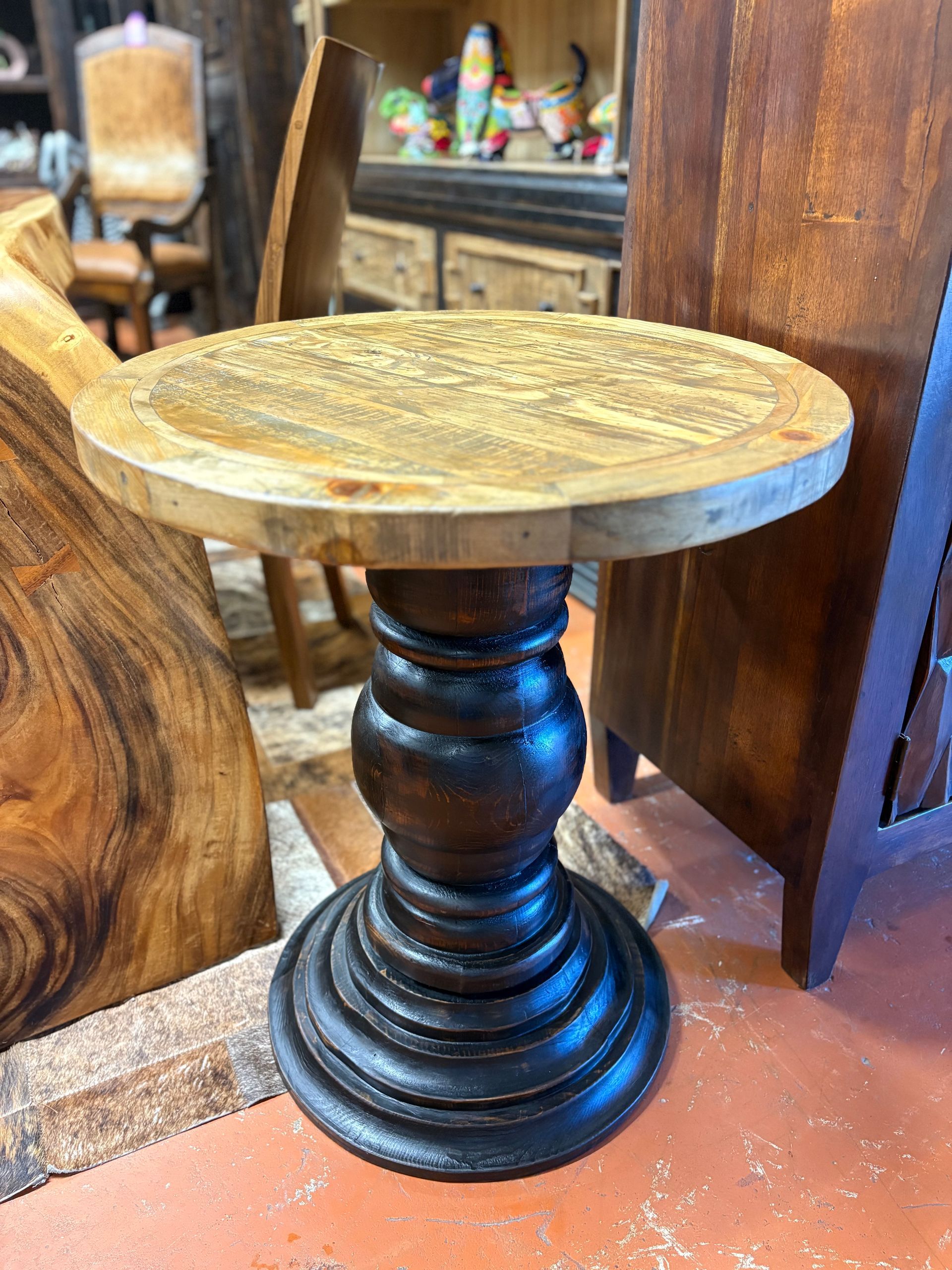 A small, round wooden side table with a light wood top and a dark, turned pedestal base, standing in a shop.