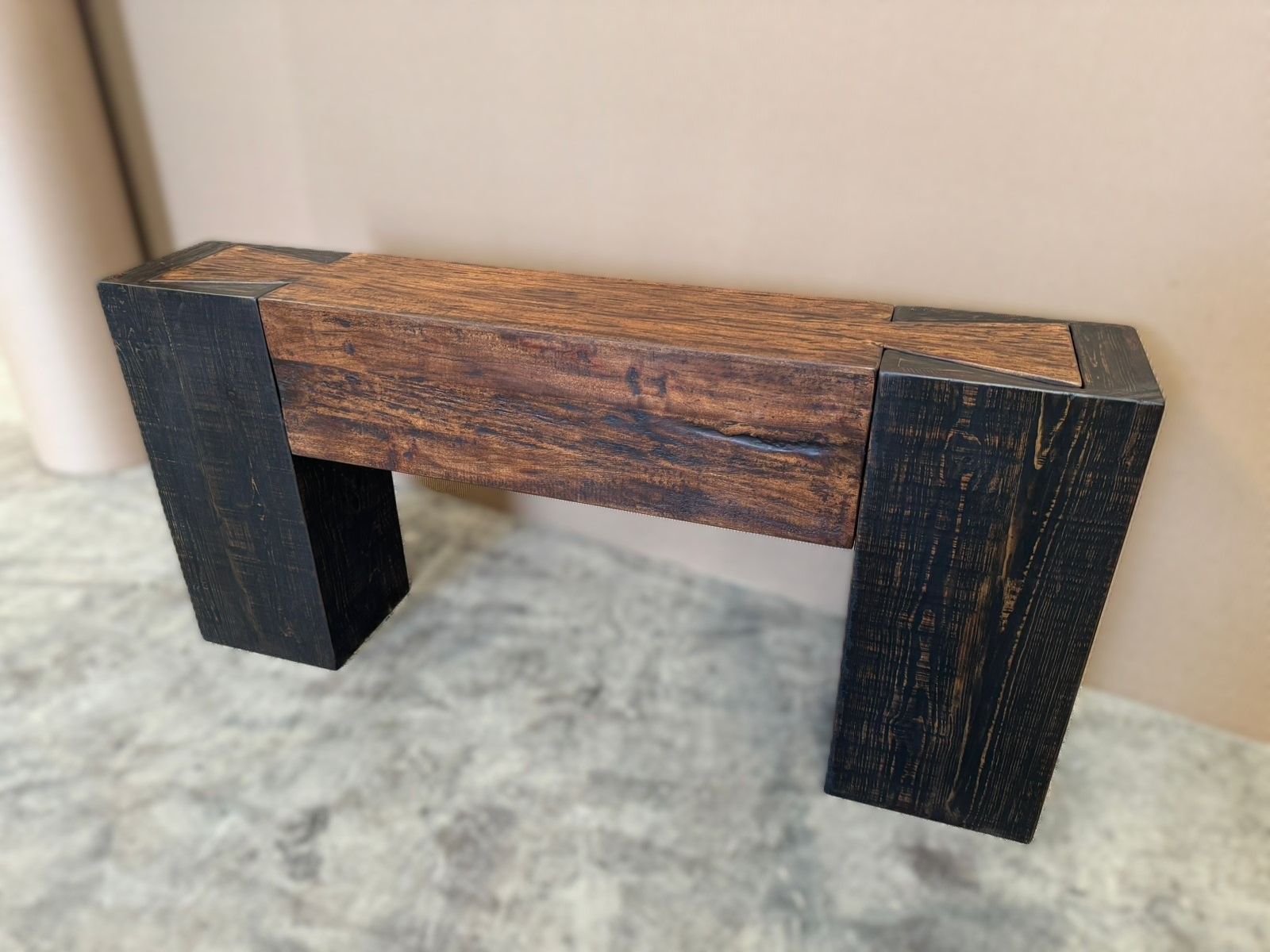 A rustic bench with a dark brown seat and black legs, sitting on a textured gray surface against a light brown wall.