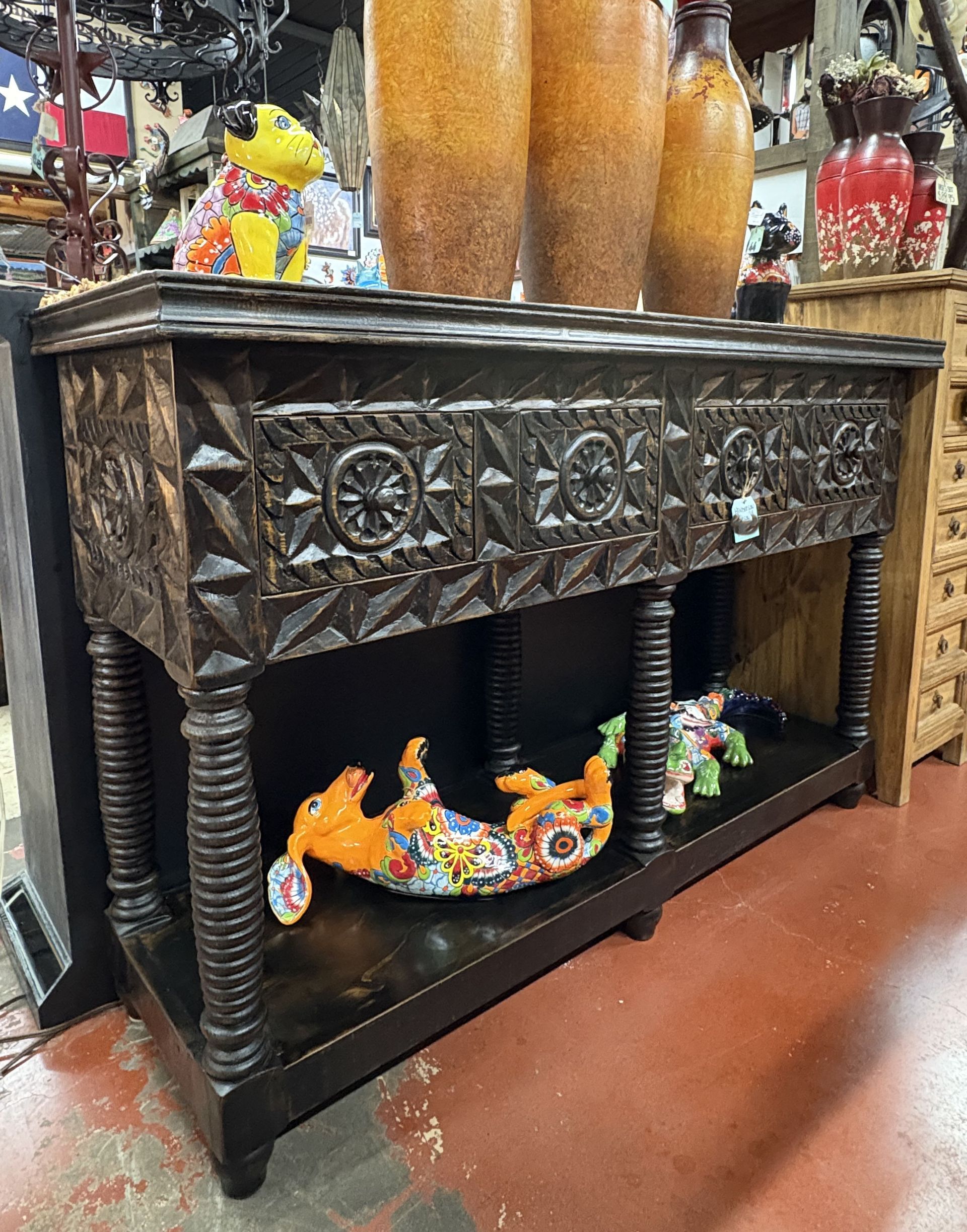 Dark brown wooden console table with carved front, two drawers, and lower shelf, displayed in a shop with decorative items on top and beneath.