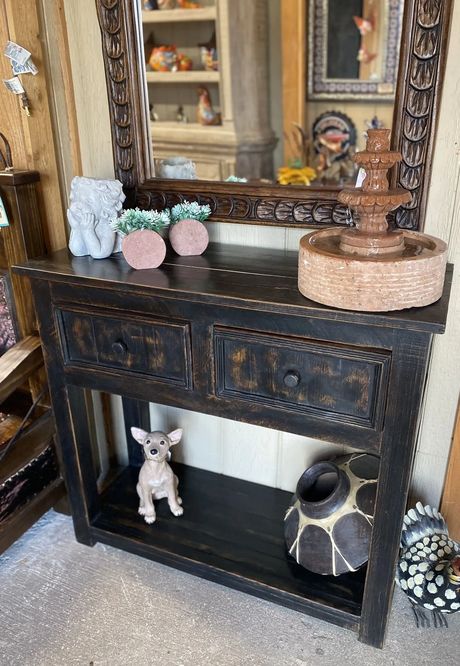 A black console table with two drawers and a shelf, displaying decor like a fountain, figurines, and pottery, reflected in a mirror.
