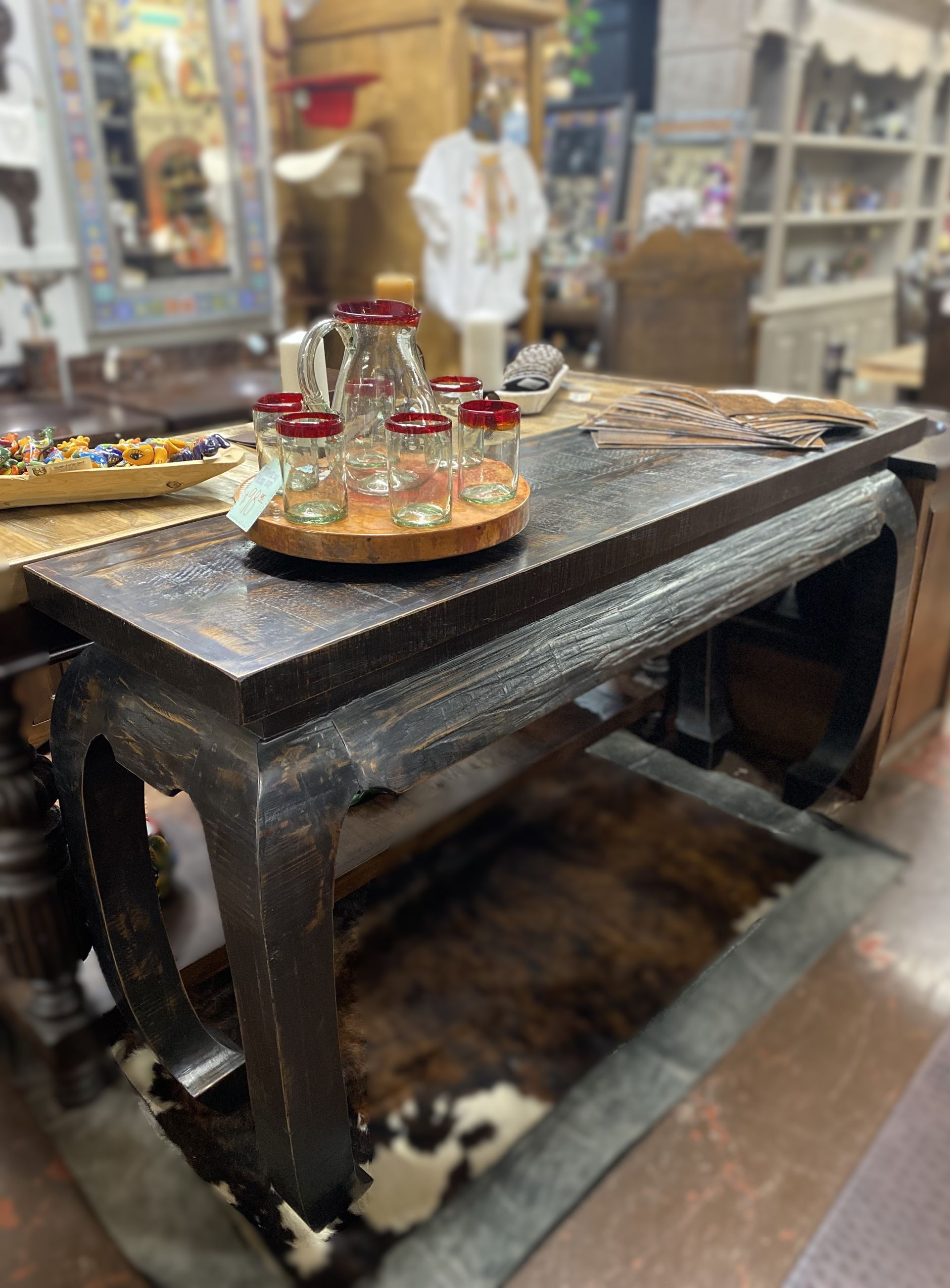 A dark, weathered coffee table with a serving tray of glassware, set in a shop. A cowhide rug is underneath.